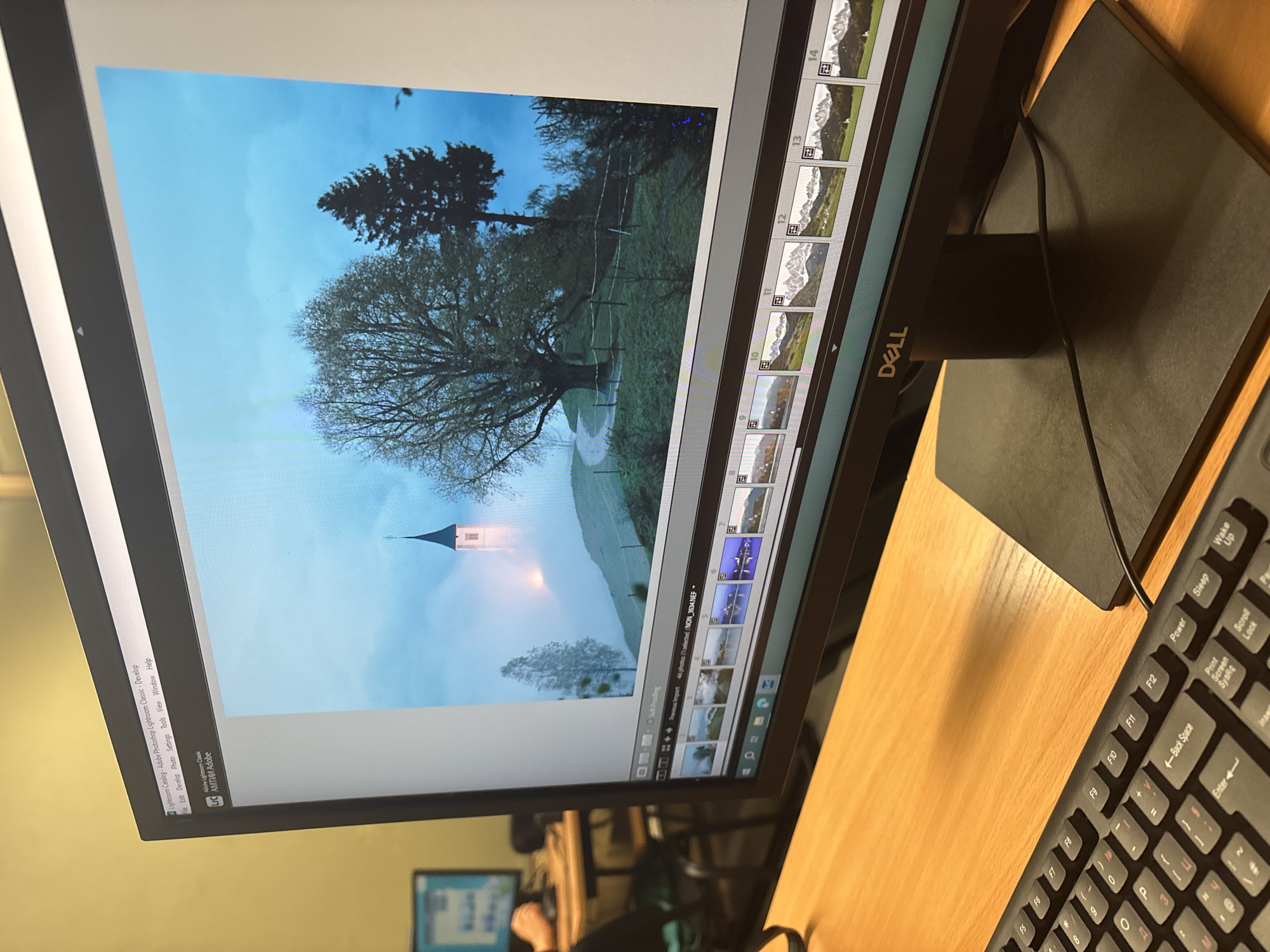 A close-up shot reveals a computer monitor displaying an image of a serene countryside scene, likely being edited in Adobe Lightroom Classic. The image features a church steeple emerging from a misty landscape, with trees in the background. A sequence of thumbnails along the right edge of the screen indicates other versions or edits of the photograph. A black Dell monitor and keyboard are visible on a light wood desk. The setting appears to be an office or workspace, with a hint of another computer visible in the background. The photograph does not have the information necessary to identify the location as Plovdiv, Bulgaria.