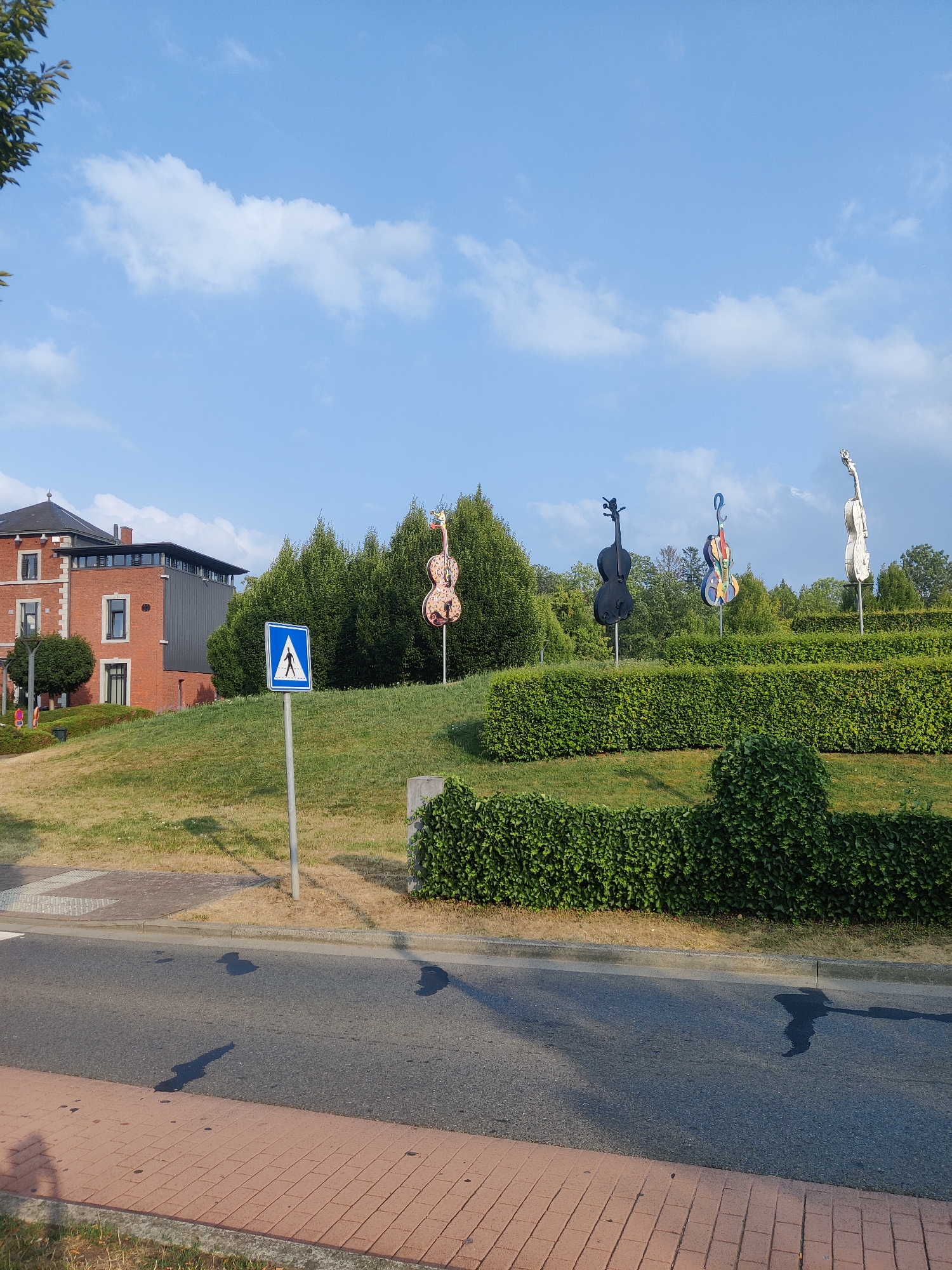 The image depicts an outdoor scene in Marche-en-Famenne, Belgium. In the foreground, a paved road runs horizontally across the bottom of the frame, with a red brick sidewalk on the left. Shadows of objects, possibly trees or structures, are cast on the road and sidewalk. Beyond the road is a grassy slope.
A pedestrian crossing sign, a blue square with a white triangle containing a white pedestrian symbol, is visible on a metal pole on the grassy slope. Behind the sign, a large building complex with red brick and dark-paneled sections is situated to the left.
Further up the grassy slope and behind the building, a line of sculpted, large-scale violin-shaped artworks are displayed. These sculptures are positioned in front of a dense line of green trees and hedges. The violin sculptures vary in color and design; one is predominantly pink and orange with intricate patterns, another is solid black, a third is multi-colored, and the furthest is a lighter, metallic color.
The sky above is blue with scattered white clouds. The overall impression is a public space with artistic installations integrated into the landscape.