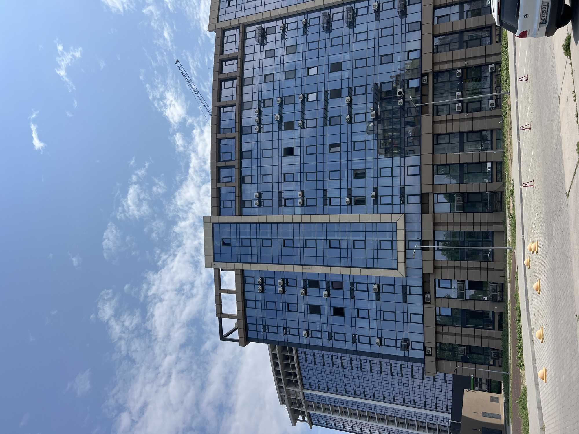 In Fontanka, Ukraine, a large, modern glass-fronted apartment building with multiple floors is visible under a bright blue sky with scattered white clouds. The building features numerous air conditioning units mounted on its exterior, reflecting the surrounding environment. A construction crane is positioned above and to the right of the building, suggesting ongoing development in the area. In the foreground, a paved parking lot with yellow parking bollards and some red parking delineators is visible. A white car with a license plate is parked in the lot. The building's facade is a combination of reflective blue glass and beige panels.