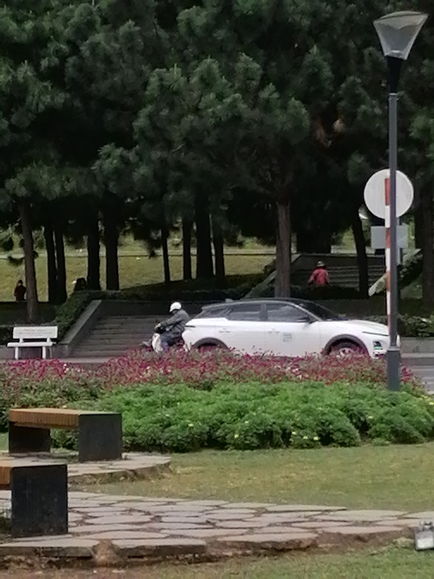 The image depicts a scene in Da Lat, Vietnam, with a focus on a white car parked next to a set of stairs. A person wearing a white helmet is riding a scooter past the car. In the background, a woman in a red outfit is visible standing near a tree line. The environment includes lush green trees, manicured bushes with red flowers, and a paved walkway. A street lamp with a circular sign is also present on the right side of the frame.