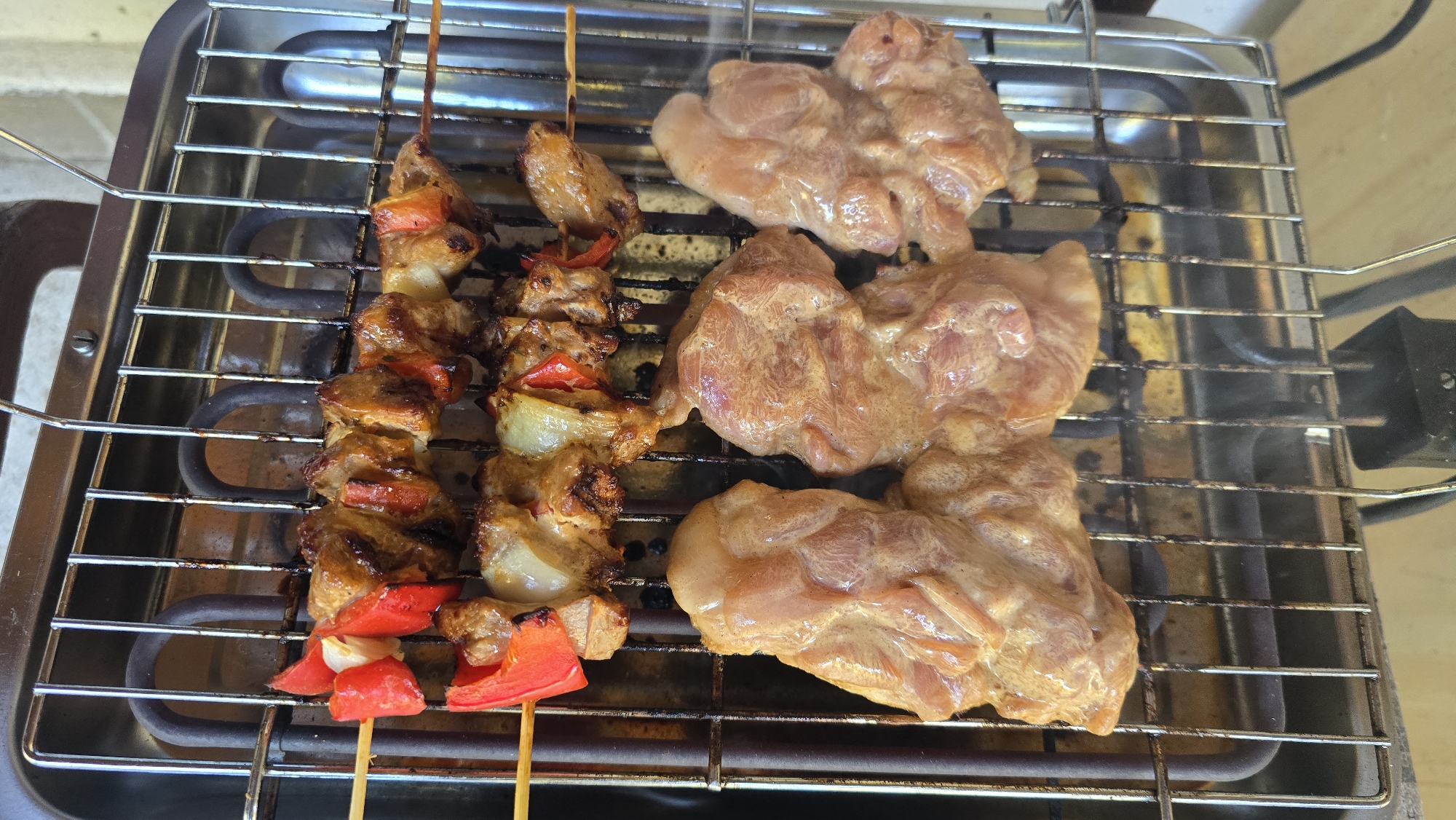 A close-up, top-down shot shows a stainless steel electric grill with various food items cooking on its grates. On the top rack, two skewers are laid out horizontally. Each skewer contains alternating pieces of browned meat, red bell pepper, and what appears to be onion. The meat on the skewers is visibly cooked and slightly charred in places. Below these skewers, on the lower grate, are three pieces of raw chicken. The chicken pieces are irregularly shaped and coated in a light-colored marinade, giving them a slightly wet and glistening appearance. The grill itself is placed on a countertop, and the heating elements are visible through the grates. The overall scene suggests food preparation, likely in a domestic setting. The image does not contain any recognizable landmarks or elements that would indicate a specific city or location like Sirishtnik, Bulgaria.
