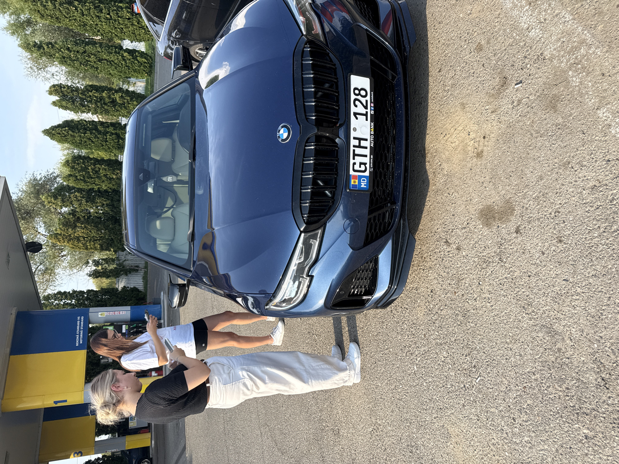 Two young women are standing next to a dark blue BMW sedan at a gas station. The woman on the left, with blonde hair tied in a bun, is wearing a black short-sleeved top and loose white pants. She is looking down at her phone, which she holds in her right hand, and has her left hand on her hip. The woman on the right, with long brown hair, is wearing a white t-shirt and black shorts. She is also looking at her phone, which she holds in her left hand. The car's license plate reads "GTH 128 MD". The gas station has yellow and blue pumps, and the scene is set outdoors with trees in the background under a clear sky. The location appears to be a gas station in the city of Hânța, Romania.