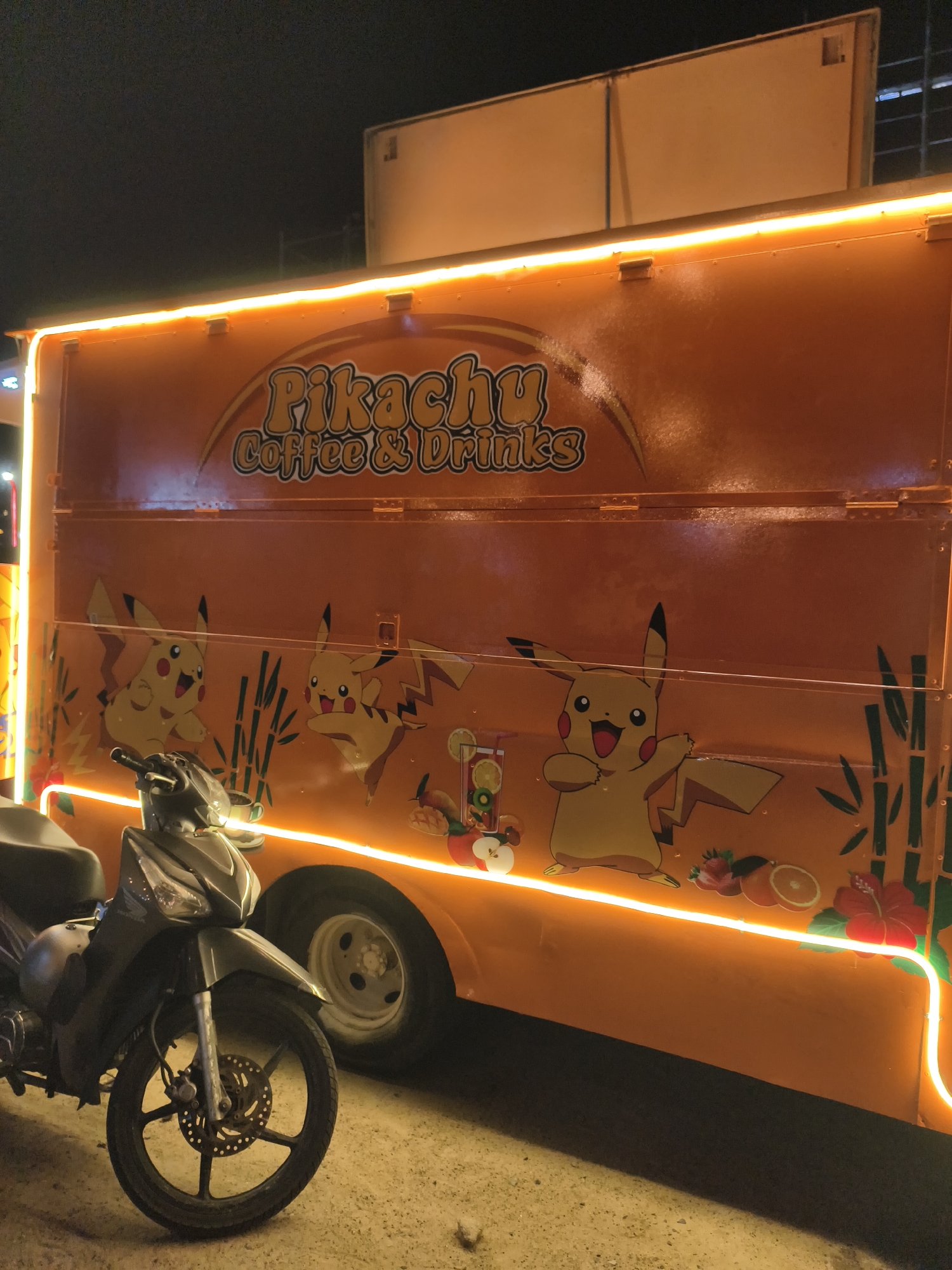 A motorcycle is parked in front of a truck with the words "Pikachu Coffee & Drinks" and Pikachu cartoon drawings on it. The truck is lit with LED strips on the top and bottom. The location appears to be in a city due to the presence of a building structure in the background. The environment is dimly lit, suggesting it might be nighttime. The city location is unknown.