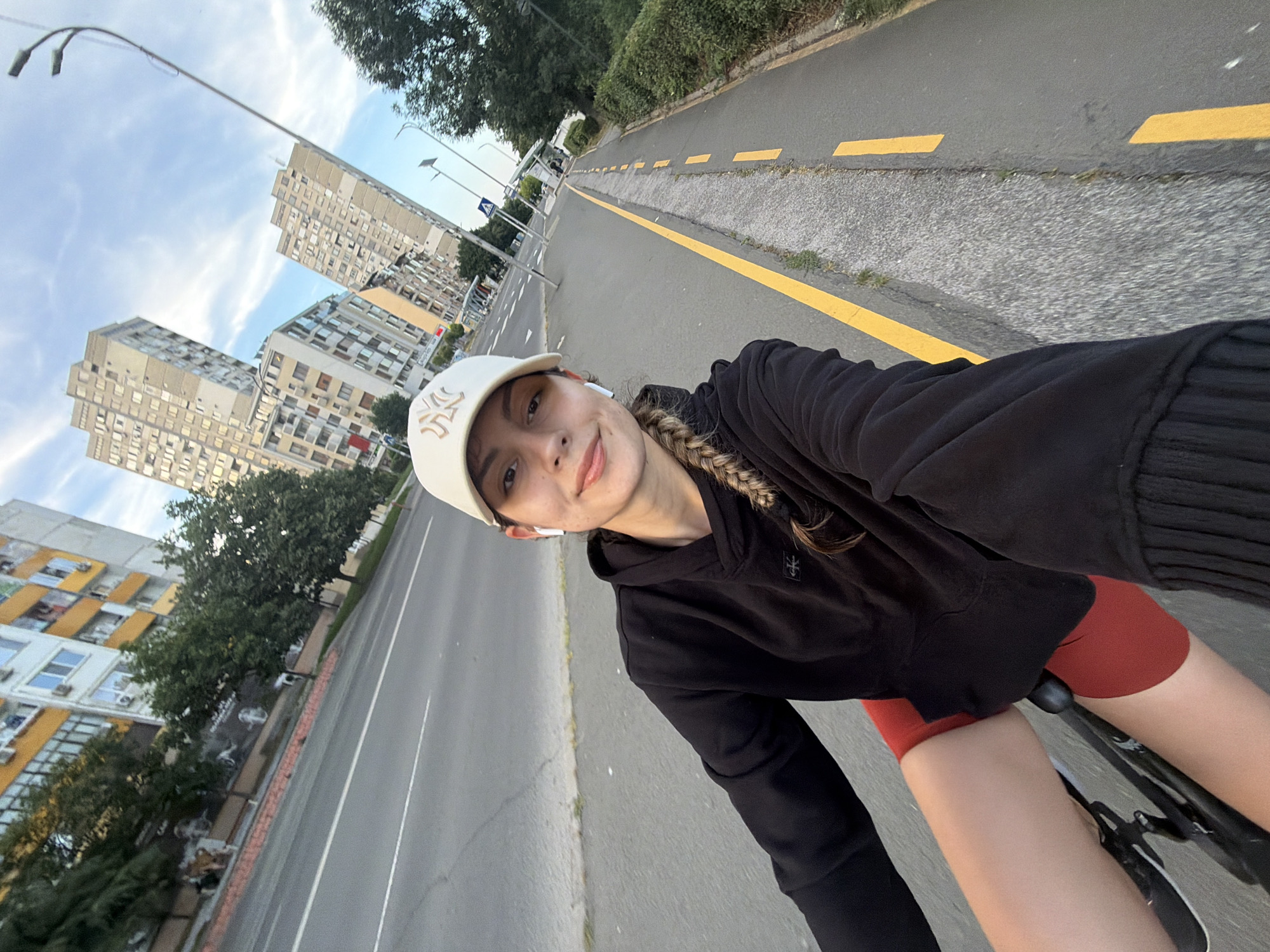 A young woman is riding a bicycle on a street in Burgas, Bulgaria, as indicated by the architecture typical of post-communist era buildings and the presence of a yellow lane divider often seen in Eastern European cities. The woman is wearing a black hoodie, red shorts, and a beige baseball cap. She has white wireless earbuds in her ears and her hair is in a braid. The background features a road, a sidewalk, several tall, multi-story buildings, and trees. The sky is partly cloudy, suggesting it is daytime. The perspective of the image is from the perspective of the cyclist, suggesting a selfie.