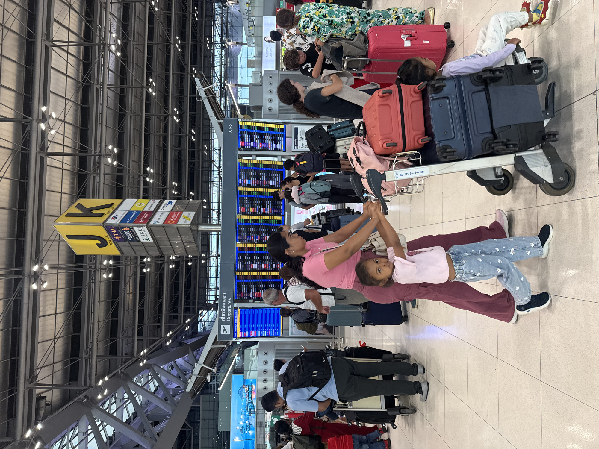 The image depicts a busy airport departure hall, likely at Suvarnabhumi Airport in Nong Prue Subdistrict, Thailand. In the foreground, a woman in maroon pants and a pink shirt pushes a luggage cart with two children. A younger girl in a pink shirt and patterned blue pants walks beside her, while a younger child sits on the cart between two pieces of luggage. Behind them, other travelers with their luggage are visible, and large departure boards display flight information overhead. The environment features a tiled floor, a high ceiling with structural beams and lighting, and prominent signage.