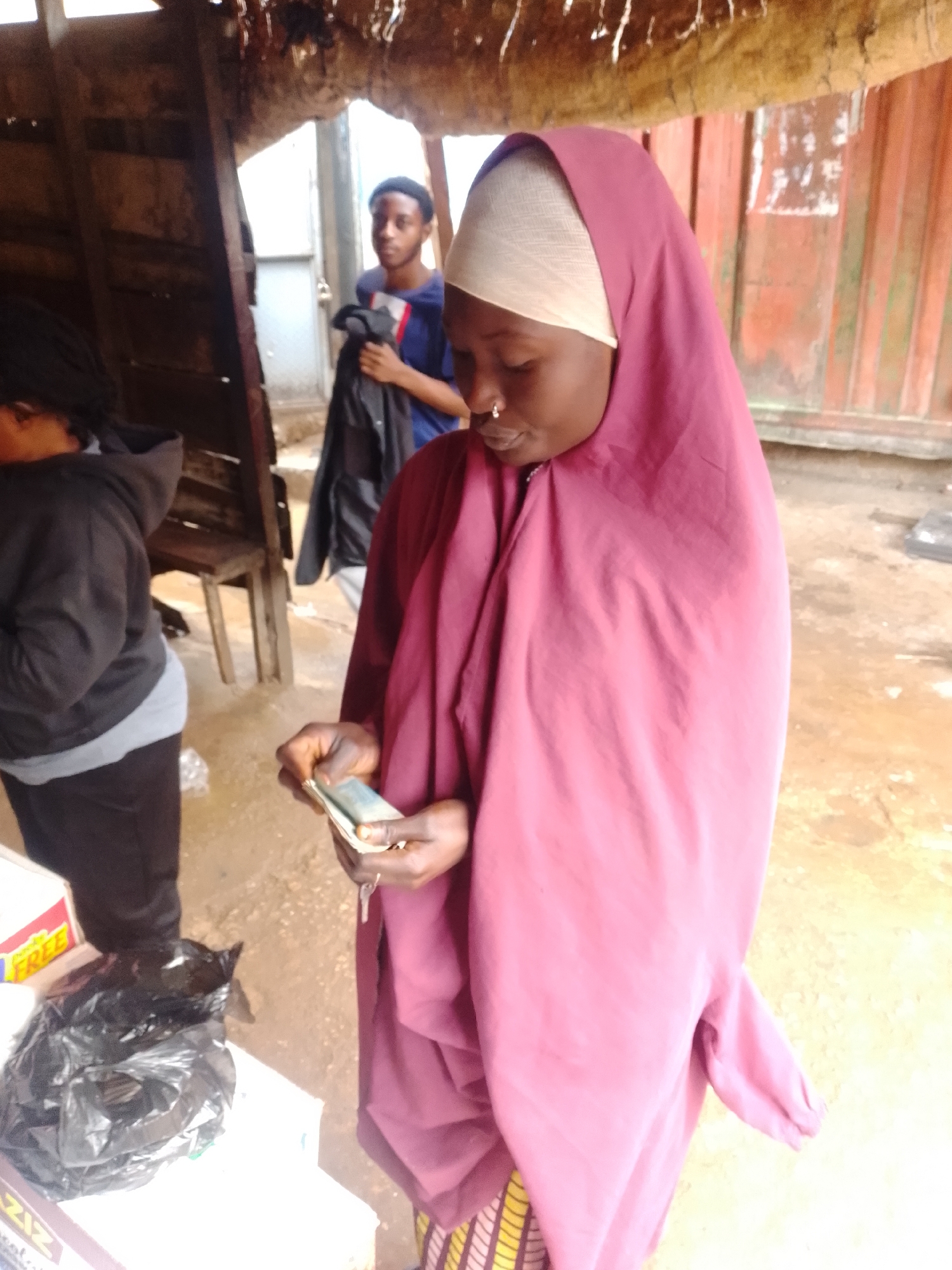 In an outdoor market setting in Unknown, Unknown, a young woman wearing a purple hijab and a tan headscarf is counting a wad of bills. She has a nose ring and is looking down at the money. Behind her, another young man in a blue shirt and black jacket stands near a wooden structure. To the left, a person in a black hoodie and gray undershirt is partially visible, facing away from the camera. In the foreground, a black plastic bag is on a white surface, next to a box with the word "FREE" visible. The ground appears to be dirt, and corrugated metal sheeting is visible in the background, suggesting a rustic or informal market stall.