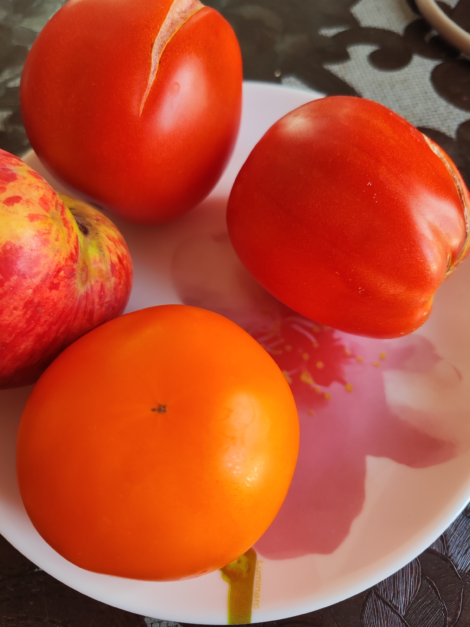 Four fruits are arranged on a white plate with a pink floral design. From left to right, top to bottom: a red apple with yellow and red streaks, a bright red tomato with a visible slit on its surface, another bright red tomato, and an orange persimmon. The plate rests on a dark, patterned surface, possibly a tablecloth. The lighting suggests an indoor setting, likely a kitchen or dining area in a residential property in Gvardeysk, Russia. There are no people or any ongoing actions depicted in the image.