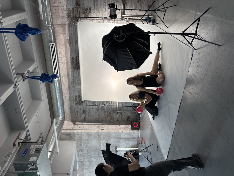 The scene depicts an indoor photography studio during an active photoshoot. The location is in a city identified as Unknown, Unknown, characterized by an industrial aesthetic with exposed brick and concrete walls, a high ceiling featuring a Carrier HVAC unit, silver air ducts, and two bundles of blue fabric suspended from structural beams. The floor is grey concrete, partially covered by a white seamless paper backdrop secured with blue tape.
Three individuals are present. On the left, a photographer with dark hair, wearing a black top and dark pants, is standing and operating a camera, aiming it towards two subjects. To the photographer's immediate right (camera-left), a large black softbox is positioned on a stand.
Two female subjects are seated on the white seamless backdrop in the center-right. The subject on the left has long blonde hair and is wearing a black short-sleeved top and black wide-leg pants. She holds a red exercise ball in her left hand and looks towards the right. The subject on the right has long brown hair and is wearing a black tank top/bodysuit with a black and white striped sock on her left foot; her left leg is extended. A second red exercise ball rests on the backdrop in front of her. She is looking upwards and slightly to the right with a faint smile.
Photography equipment includes a large black parabolic softbox labeled "L-900 Smilimg" on a stand in front of the subjects, and a studio strobe head on a stand to the far right. A black speaker with a circular red illuminated ring is visible on the floor against the concrete wall on the left, connected by an orange extension cord.