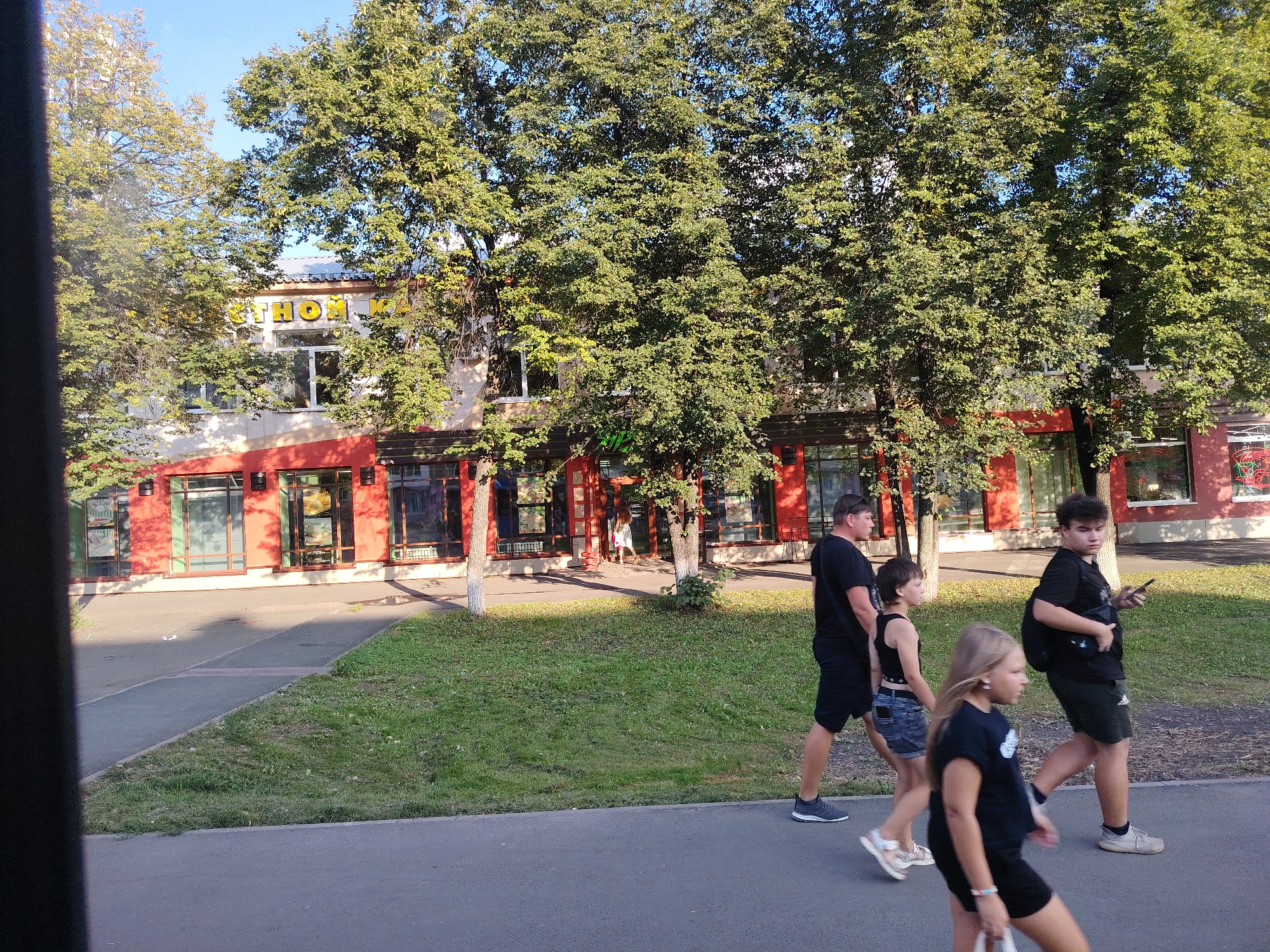 The image shows a street scene in Kemerovo, Russia. A group of people are walking on a sidewalk in front of a building with a red facade and large windows. The building appears to be a commercial establishment, with the letters "THOM" visible on its facade in yellow. Trees with green foliage line the sidewalk and partially obscure the building. The ground is paved with asphalt and concrete, with a strip of grass between the sidewalk and the building. The sky is clear and blue, indicating a sunny day. The people in the image are dressed in casual clothing, and some are carrying bags. The overall scene suggests a typical day in a city.