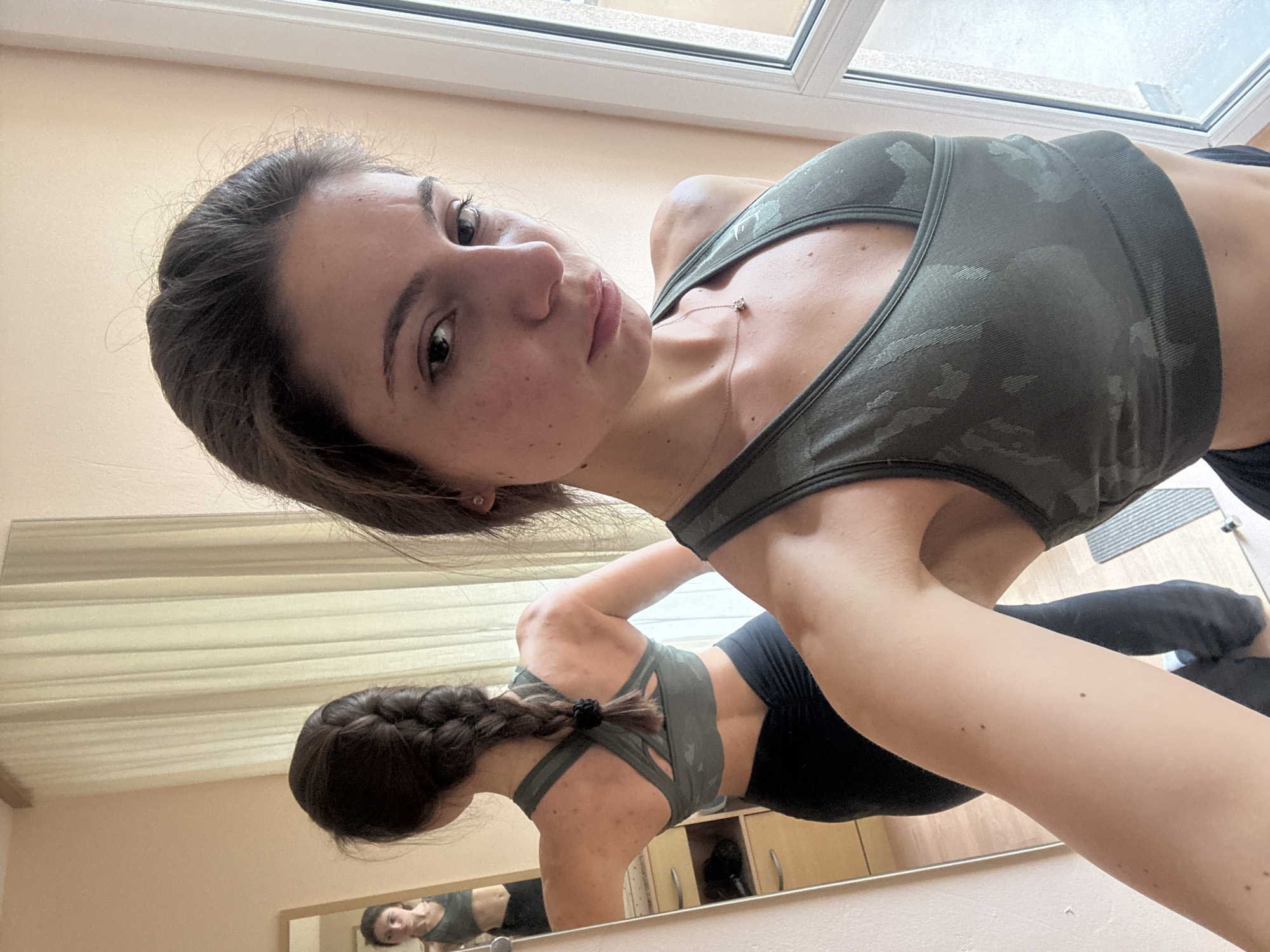 The image depicts a woman in a green camouflage sports bra and black shorts, seemingly posing for a selfie in a room with a large mirror and a window. She is positioned at an angle, looking towards the camera. Her dark hair is partially pulled back, and her reflection is visible in the mirror. The room appears to be a gym or studio, given the exercise attire and mirrored wall. Cabinets are visible along the wall beneath the mirror. The window is towards the top of the frame, suggesting a bright, possibly daytime setting. No specific indicators suggest Varna, Bulgaria.