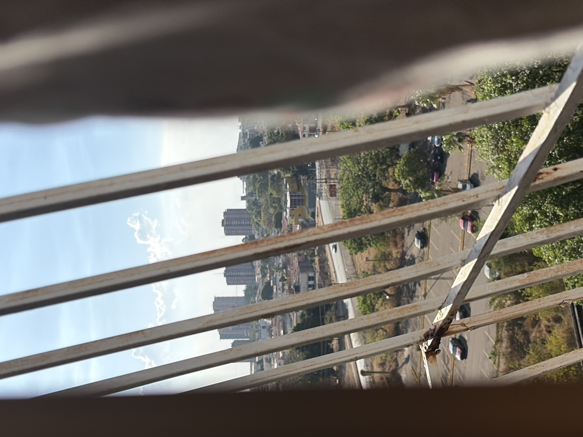 The view from behind a weathered white metal balcony shows the city of Ciudad Guayana, Venezuela. In the distance, there are several high-rise apartment buildings. A road with parked cars runs parallel to the balcony, separated by trees and vegetation. The sky is visible through the balcony railings and is mostly blue with some clouds. The balcony appears to be older, with rust visible on the metal structure.