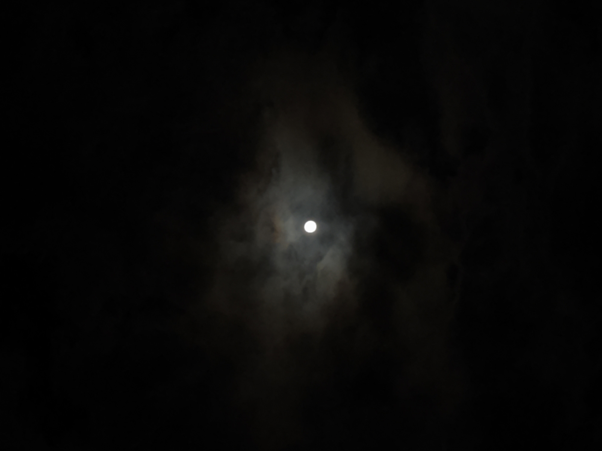 A dark, nighttime scene with the moon partially obscured by clouds. The moon is a bright, circular object in the center of the frame. The surrounding clouds are varying shades of gray, creating a hazy, atmospheric effect. The rest of the scene is enveloped in darkness. I cannot determine the specific location in the city of Kozloduy, Bulgaria.