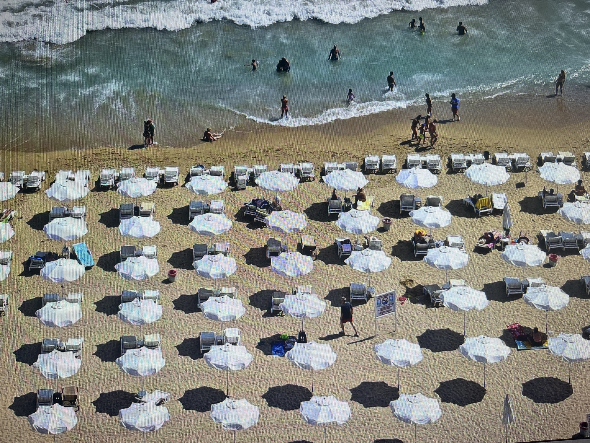 This high-angle, slightly pixelated image depicts a vibrant beach scene in Varna, Bulgaria, under bright daytime conditions. The top third of the frame shows the clear, turquoise sea with white-foaming waves, where numerous individuals of various ages are swimming, wading, and standing in the shallows. The sandy shoreline features a few people closer to the water, including what appears to be a couple embracing and another person relaxing on the sand.
The majority of the beach is covered by a grid-like arrangement of white beach umbrellas, each accompanied by one or two white or light-colored sun loungers. These umbrellas cast prominent, dark shadows onto the golden sand, suggesting a high sun. Scattered throughout the umbrella section are several beachgoers: some are seated or lying on loungers, others are walking along the sandy paths between the rows, and one person is attending to a blue mat.
Notable objects include several red waste bins and at least two signs. One sign, situated amidst the umbrellas, features a red circle with a diagonal line, indicative of a prohibition or warning, but the text is not legible. Another smaller sign is visible further left. The overall scene portrays a typical, bustling summer day at a popular beach resort, filled with leisure and recreational activities.
