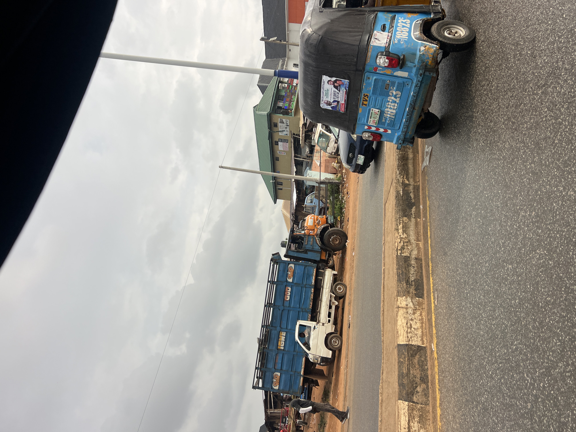 The image depicts a street scene in Okpanam, Nigeria, under an overcast sky. In the foreground, a blue auto-rickshaw (Keke Napep) is parked on the right side of the road. It has a poster on its rear advertising a "Dawn to Dusk" event, featuring images of two individuals. The rickshaw bears license plates that read "CYV 819SZ" and "18823". A dark blue car is visible behind the auto-rickshaw.
Further back, a large blue truck with the words "IGWE GOD" painted on its side is parked on the left side of the road, angled towards the street. A small white truck is positioned in front of it. A man in a dark t-shirt and grey pants is walking away from the trucks towards the left edge of the frame. Buildings, some with green roofs, line the background, suggesting a commercial or residential area. The road surface appears to be asphalt with a yellow line marking the lane. A concrete curb with black and white markings separates the road from a dirt area where the trucks are parked. The overall impression is of a typical street scene in a Nigerian town, possibly with market activity or transport hubs.