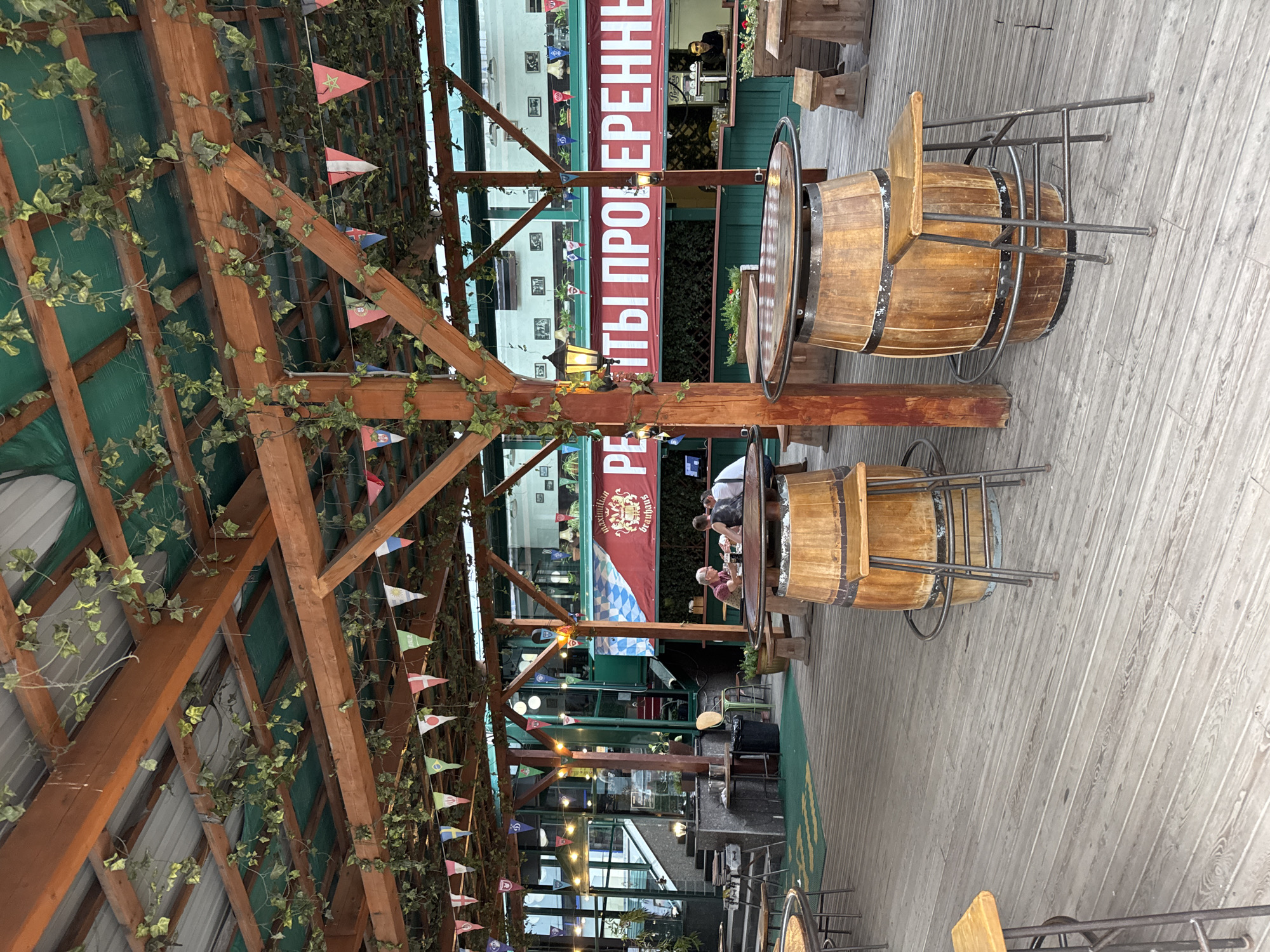 The image depicts an outdoor seating area of a restaurant or beer garden in Saint Petersburg, Russia. The scene features wooden decking and a covered structure supported by wooden beams and posts, adorned with artificial ivy and small triangular flags of various countries. Several barrel-shaped tables with attached stools are arranged on the deck, and in the background, a large red banner with white Cyrillic text is visible. A few patrons are seated at a table in the middle ground, engaged in conversation. The overall ambiance suggests a casual dining or drinking establishment.