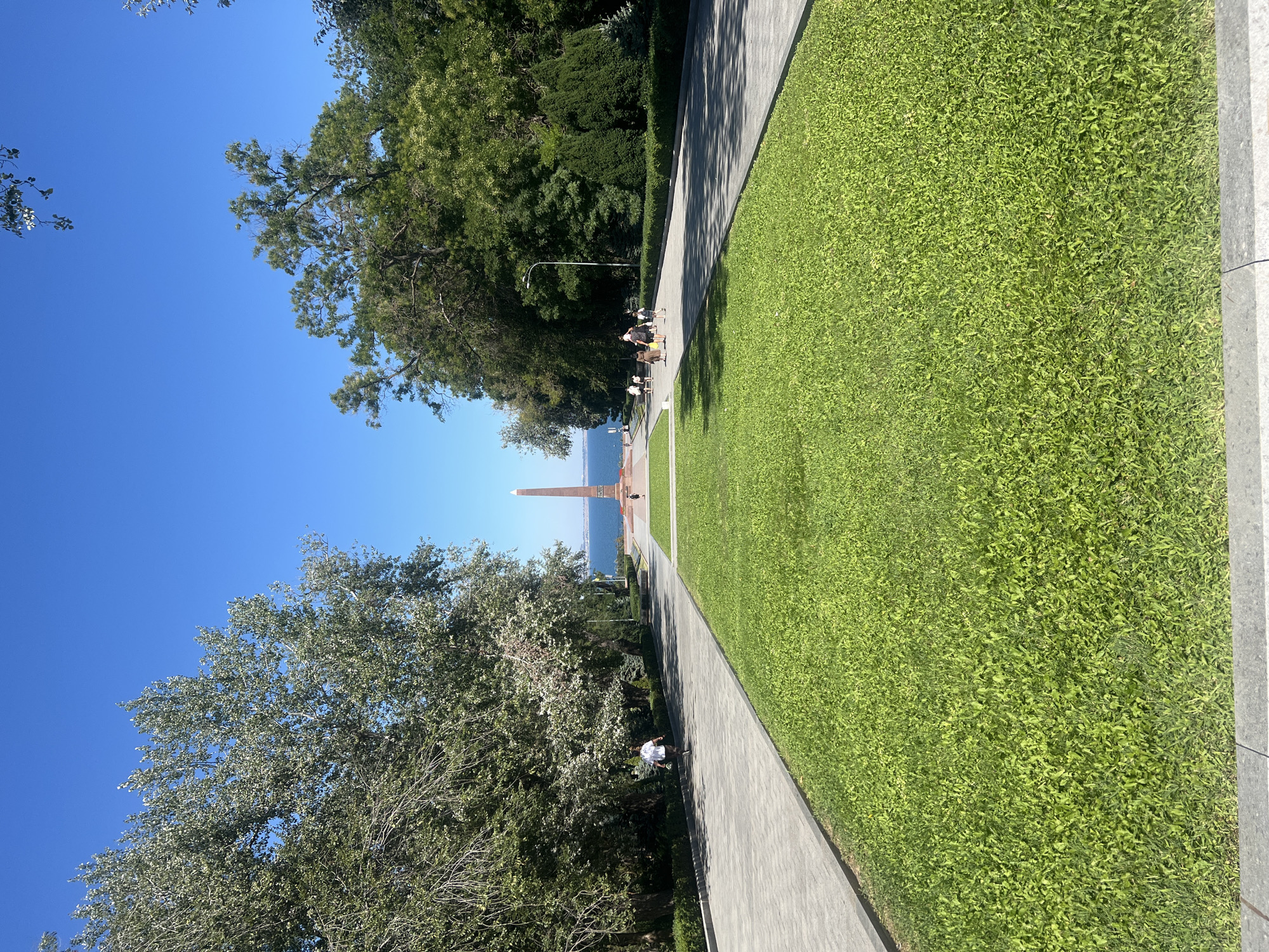 This is a wide-angle, eye-level shot of a memorial park in Odesa, Ukraine. The scene is dominated by a bright, clear blue sky and lush green grass. In the foreground, a wide, paved walkway stretches into the distance, bordered by a neatly manicured lawn. To the left and right of the walkway, mature trees with full canopies line the path, casting dappled shadows. A lone person is walking away from the camera on the left-hand path.
Further into the distance, a tall, slender obelisk monument stands prominently in the center of the frame, positioned on a raised circular base. Beyond the monument, a body of water, likely the Black Sea, is visible under the clear sky. Several people are walking or standing near the monument in the middle ground. The overall impression is one of a peaceful, well-maintained public space on a sunny day.