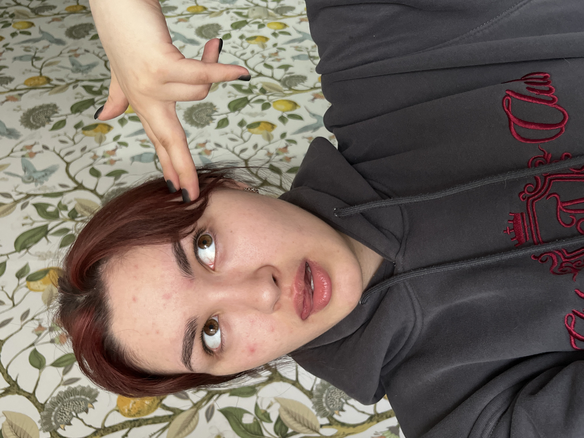 A young woman is holding her hand to her head in a gun shape, with black nail polish. She has brown hair and is wearing a gray hooded sweatshirt with red embroidery. The background is a floral print. No information relating to Neftekamsk, Russia can be found in the image.