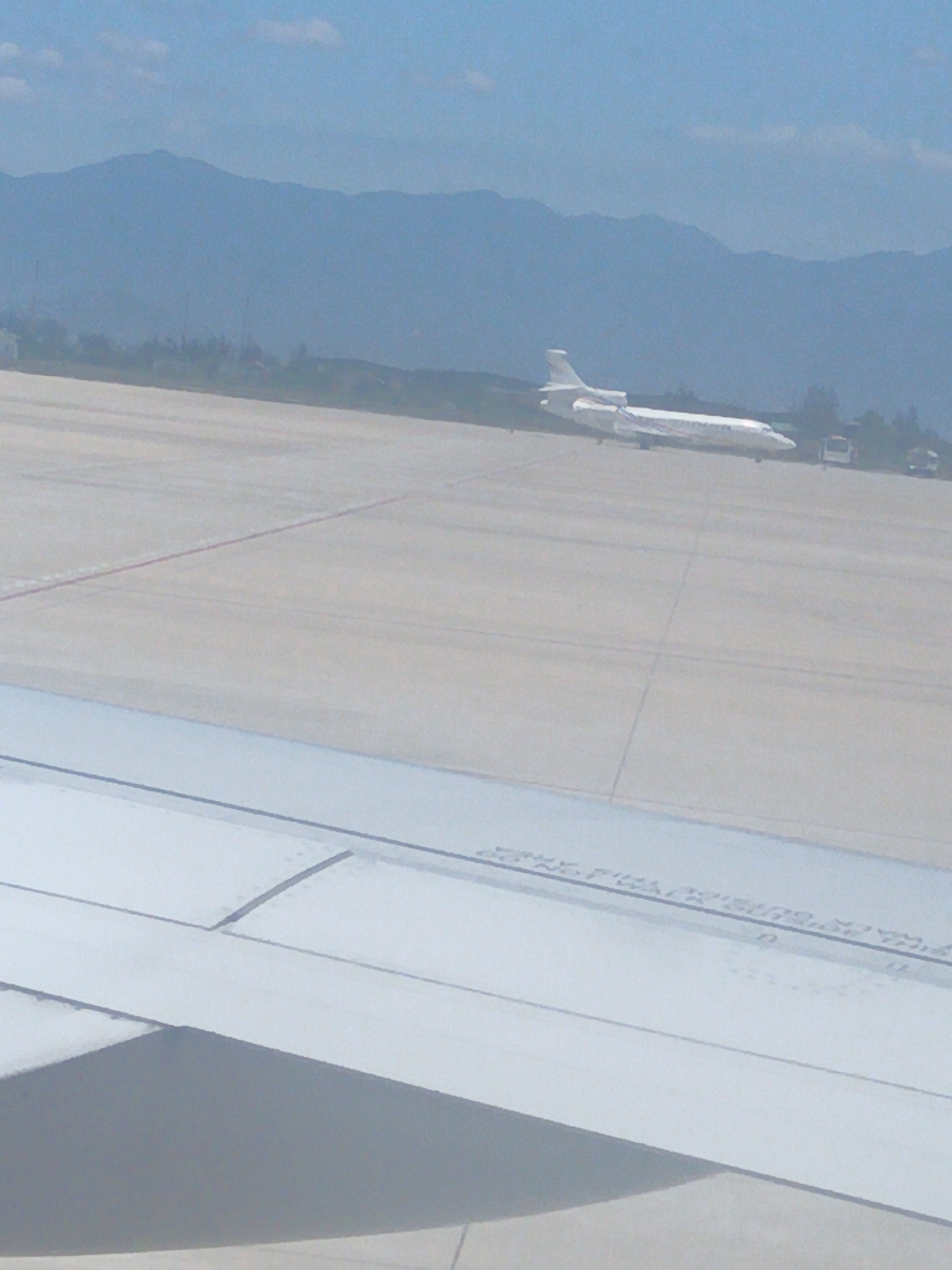The image shows an airplane on a tarmac with mountains in the background. The airplane is white with a blue stripe. There are also two buses visible on the tarmac. The sky is blue with some clouds. The image appears to be taken from an airplane window, as a portion of an airplane wing is visible in the foreground. The scene is likely at an airport in Bắc Cam Ranh, Vietnam, given the presence of mountains that are characteristic of the region. The lighting suggests it is daytime. The focus is on the airplane and the surrounding airport environment.