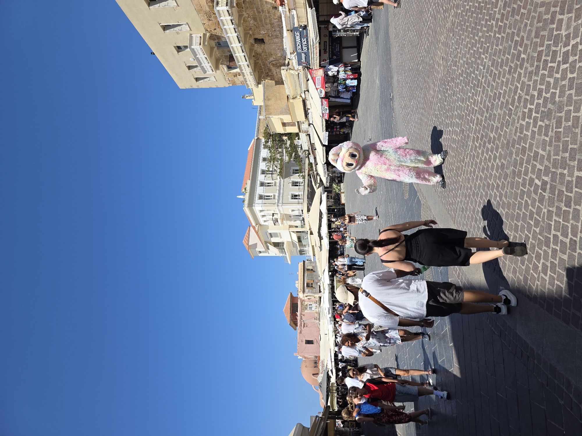 In Chania, Greece, on a clear, sunny day, a bustling pedestrian street is depicted. The foreground is dominated by a cobblestone pavement casting long shadows. On the right, a person in a fluffy, multi-colored costume, resembling a cartoon character with a large head and small eyes, waves to unseen observers. Further back, several people walk along the street, some in groups and others singly.
The street is lined with shops and stalls, many with awnings providing shade. Buildings with a light-colored facade and white balconies are visible in the background. One building on the right has a prominent sign reading "EXCHANGE OFFICE." To the left, a pink dome structure is partially visible. The sky is a vibrant, cloudless blue. The overall scene conveys a lively, tourist-oriented atmosphere.