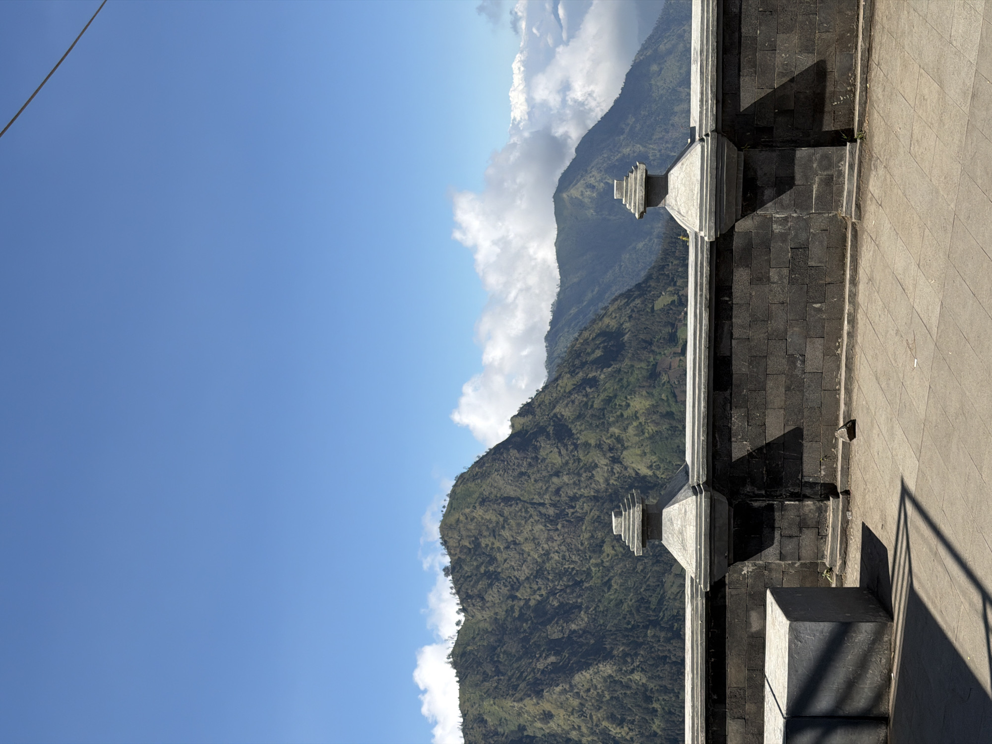 The image depicts a scenic view from a walled overlook in Cemorolawang, Indonesia. In the foreground, a dark stone wall with decorative pillars forms a barrier. To the left, two large, grey, concrete cube-shaped structures are visible, casting long shadows. The ground is paved with square tiles. Beyond the wall, a vast mountain range dominates the background. The mountains are covered in dense green vegetation, with some areas appearing rocky and barren. White clouds are scattered across a clear blue sky, with a thin wire or cable visible in the upper right corner. The overall scene suggests a viewpoint offering a panoramic vista of the natural landscape surrounding the town.