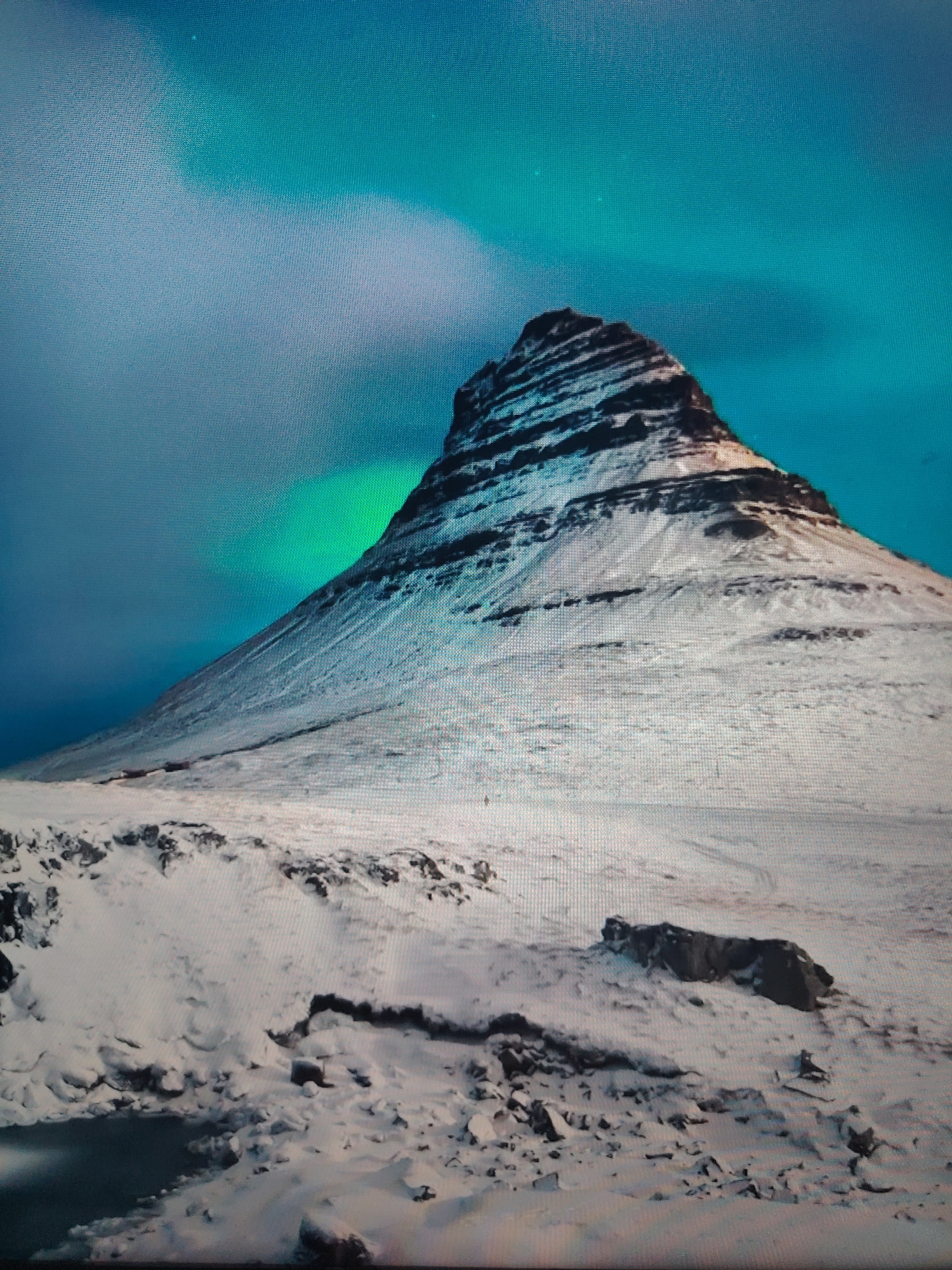The image shows a mountain covered in snow under a sky with aurora borealis. The mountain is cone-shaped with horizontal stripes of dark rock interspersed with snow. The foreground is a snow-covered plain with rocks and some darker patches, possibly indicating water or bare ground.