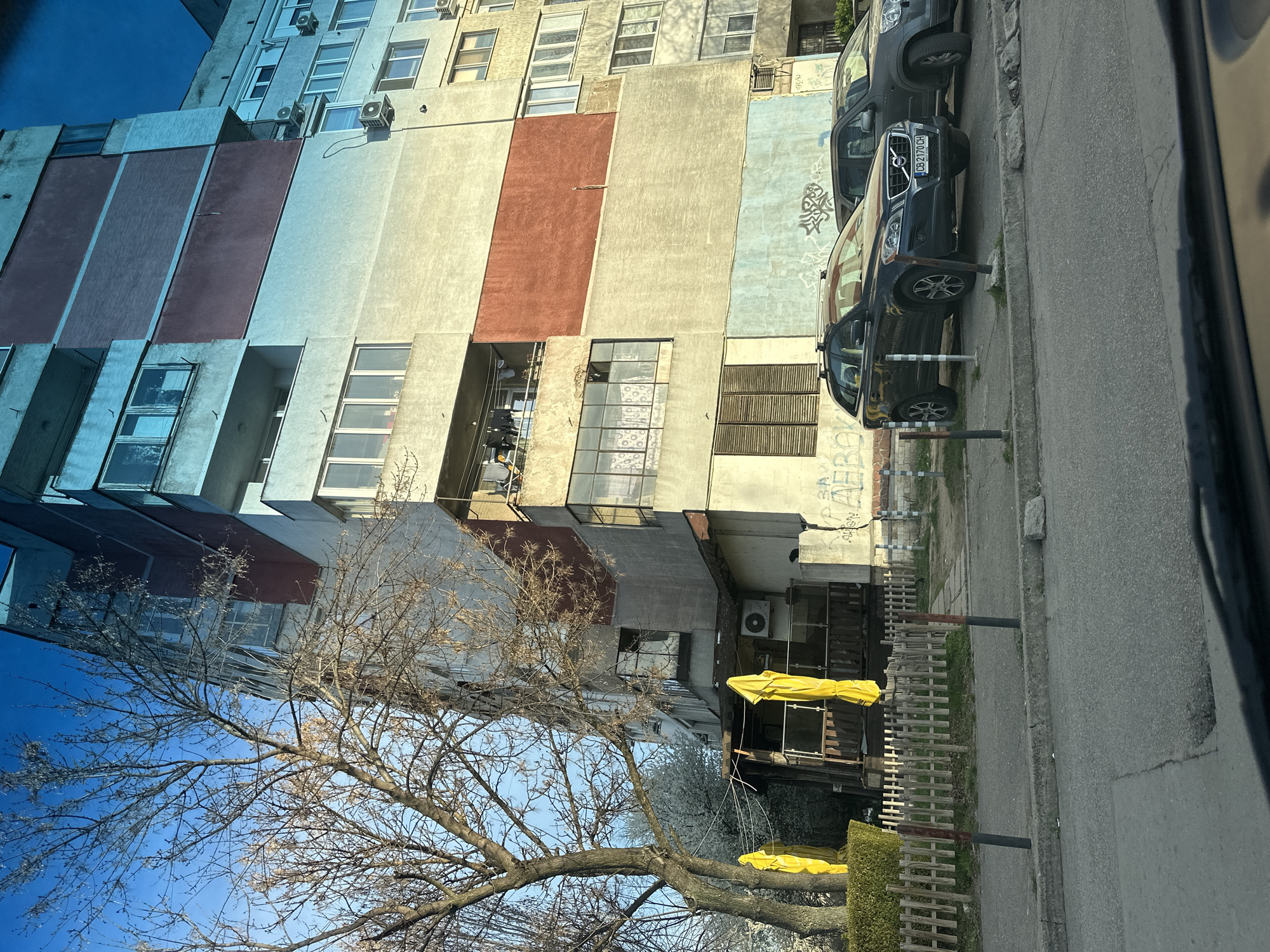 The image shows a building in Sofia, Bulgaria. The building is a multi-story apartment complex with a faded, multi-colored facade. Adjacent to the building is a sidewalk with cars parked on it. A bare tree in the foreground partially obscures the view of the building. The sky is blue, and the light suggests it is daytime. Some building features include air conditioning units, windows, and graffiti. The license plate number is CB2170CH.