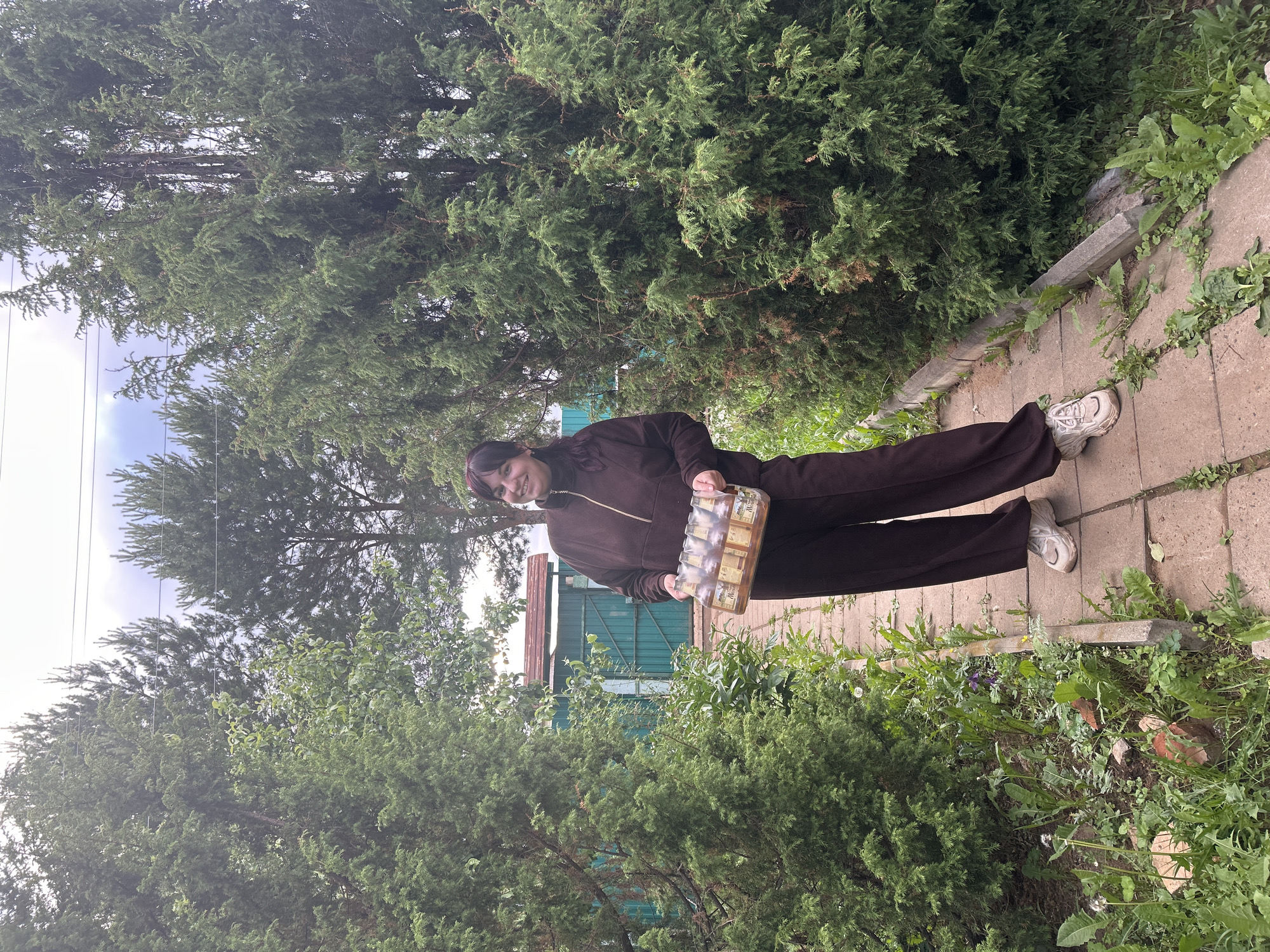 The image displays an adult female standing on a paved pathway, bordered by dense evergreen coniferous trees, within a rural or suburban garden setting in the city of Фоки, Russia. The female, possessing dark reddish-brown, shoulder-length hair with bangs, is smiling and looking directly towards the camera. She is attired in a dark brown quarter-zip tracksuit, consisting of a long-sleeved top and matching loose-fitting pants, and light-colored chunky sneakers. She is holding a clear plastic carrier containing six uniform small bottles of a light-brown liquid, each bottle featuring a label with a prominent 'M' logo.
The pathway is composed of rectangular concrete pavers, with weeds and grass visibly growing between the blocks and along the edges. Evergreen trees densely line both sides of the path, creating a verdant corridor. In the background to the left, a light blue-green metal fence or gate is visible, along with a partially obscured structure featuring a reddish-brown roof, likely a shed. Taller pine trees and additional deciduous foliage are present in the background. Thin utility lines are discernible across the upper portion of the frame, set against an overcast, light grey sky. The ground level surrounding the pathway is covered with green vegetation, including various weeds and dandelions.