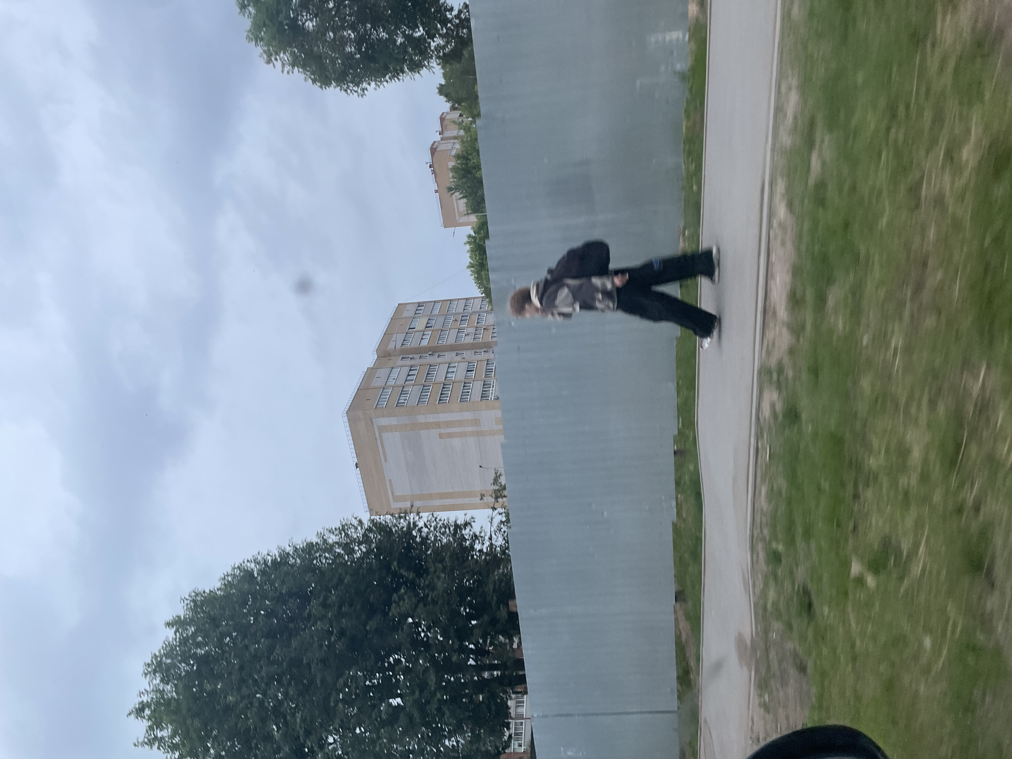 This image depicts an urban scene in Votkinsk, Russia, captured from a moving perspective on an overcast day.
In the midground, a single individual is walking from left to right on an asphalt sidewalk. The person has medium-length, curly light brown hair, is wearing a multi-toned patterned jacket (appears grey, white, black), dark trousers, and carries a black backpack. They are wearing light-soled athletic shoes, visible on the left foot. The individual exhibits motion blur, indicating movement relative to the camera.
Immediately behind the sidewalk and the walking person is a tall, light gray corrugated metal fence, appearing to be a construction barrier or property boundary.
In the background, behind the fence and trees, multiple buildings are visible. On the left, a large, multi-story residential building with a light beige exterior and a distinct vertical light gray panel design occupies the mid-background, featuring numerous windows. Further right and slightly behind, another building with a reddish-brown roof can be seen amidst dense green deciduous trees. The left side of the scene is dominated by a large, full green tree.
The sky is uniformly overcast with light gray clouds. The immediate foreground, on the left, shows a partial black car mirror, and below the sidewalk, a strip of green grass is visible. The overall scene conveys a functional, everyday urban environment under a diffused light.