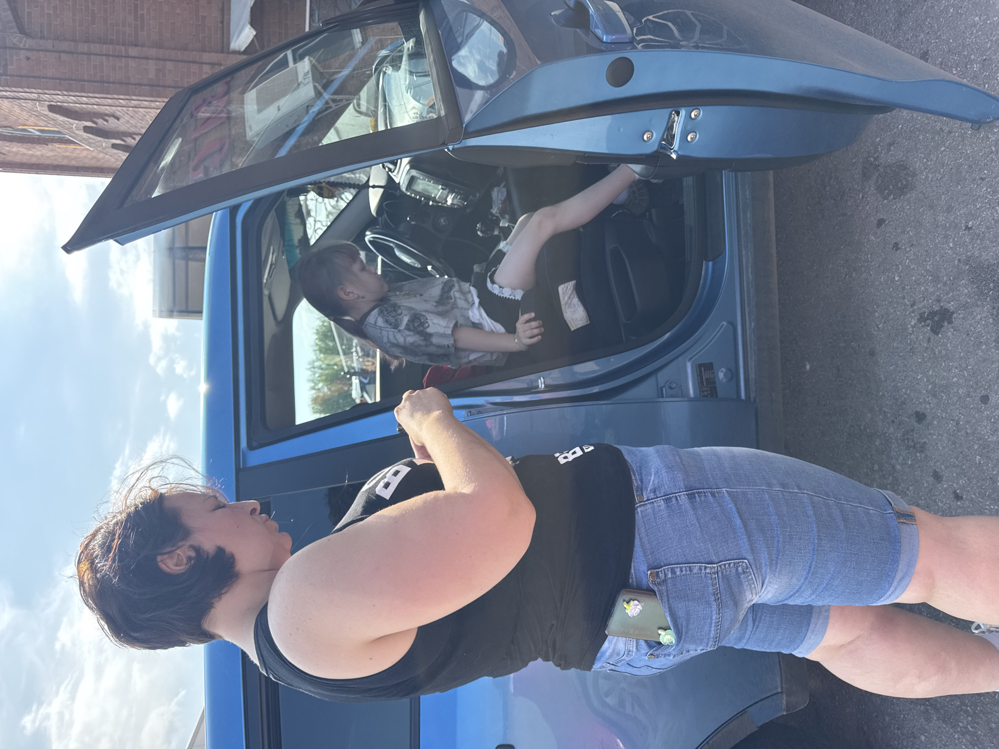 The image shows two people, a woman and a young child, in an outdoor setting. The woman is standing next to a blue car, with the driver's side door open. She is wearing a black t-shirt and denim shorts. The child is sitting in the driver's seat of the car, with their legs extended out of the door. The child is wearing a patterned t-shirt and dark shorts. The scene appears to be during the daytime, with a bright sky and some clouds visible. The background shows a brick building and some trees. There is a phone with a cartoon charm in the woman's shorts pocket. The overall mood seems casual and relaxed.