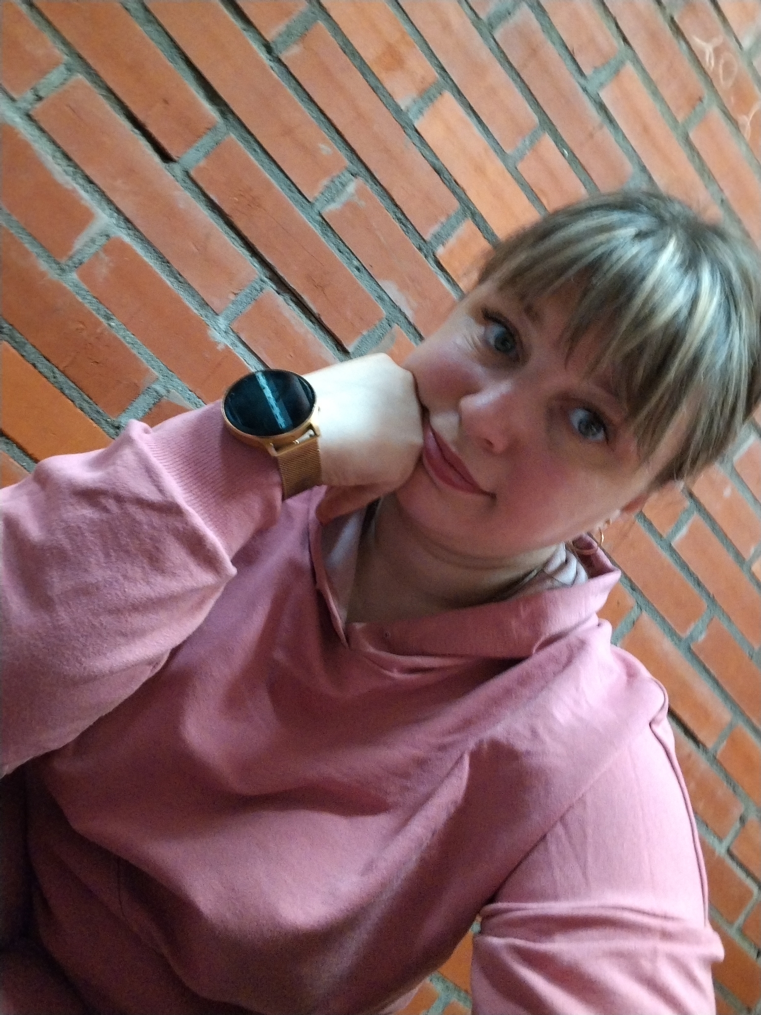 A woman poses for a close-up selfie against a reddish-orange brick wall, in Шебунино, Russia. She appears to be in her 20s or 30s, with light brown hair that has blonde highlights and is styled with bangs. Her blue eyes look directly at the camera, and she has a calm, slightly smiling expression.
She is wearing a pink hooded sweatshirt (hoodie), and her left hand is raised, with her chin resting on it. A gold-toned smartwatch with a dark, reflective screen and a gold mesh band is visible on her left wrist.
The brick wall behind her is composed of rectangular bricks laid in a diagonal pattern, separated by grey mortar. The bricks show some signs of wear and slight discoloration. In the upper right section of the wall, faint, light-colored markings or graffiti are visible, including some indiscernible letters and shapes. The lighting suggests it is daytime.