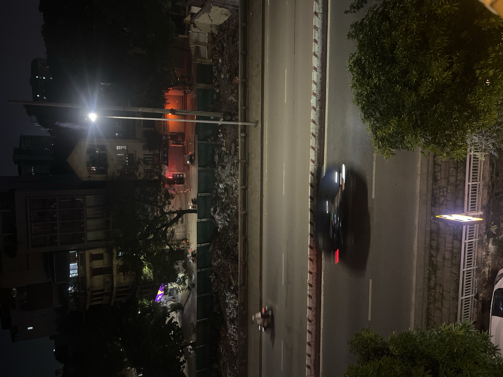 The image depicts a nighttime scene in Hanoi, Vietnam, likely along a multi-lane road. In the foreground, lush green foliage from trees obscures the lower portion of the frame, with a white railing and a lit sign visible below. The middle ground shows a busy street with a car and several motorcycles in motion, evidenced by motion blur. The road is divided by white lines, and a concrete barrier with red and white markings separates it from the area above.
Beyond the road, a construction site or demolition area is visible, characterized by a pile of debris, rubble, and a green metal barrier. Several buildings are situated behind this area, some appearing older with balconies and lit windows, while others are modern high-rises with illuminated windows. A prominent streetlamp with a bright light casts a glow on the scene. The overall atmosphere is urban and active, even at night, with the presence of moving vehicles and illuminated structures. The style of the buildings and the vehicles, particularly the motorcycles, suggest an Asian urban environment.
