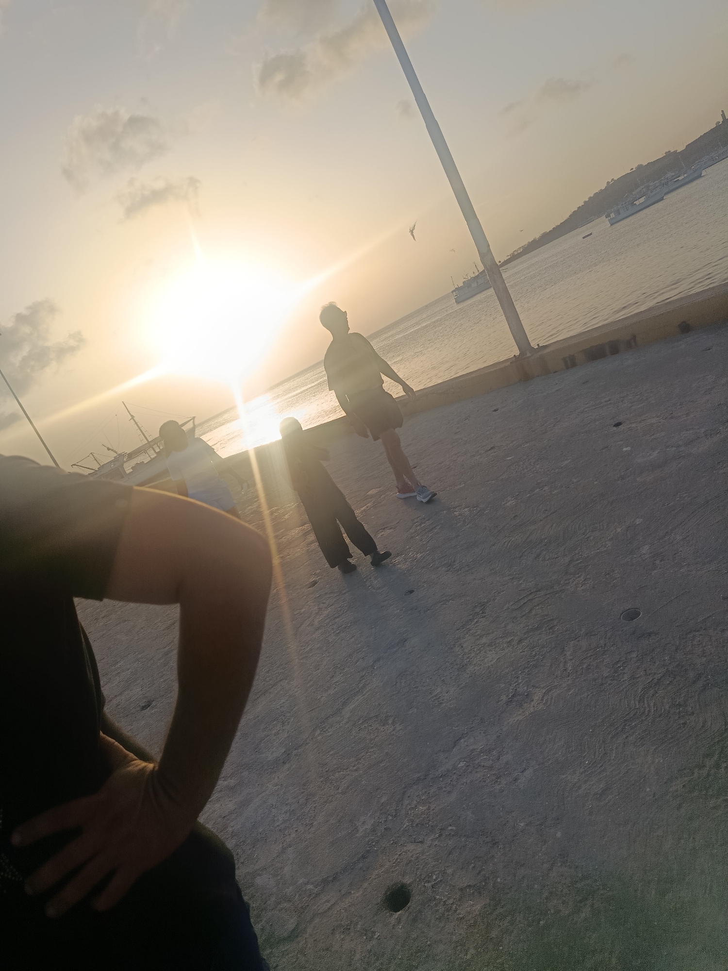 The image captures a sunset scene at the Muelle de Pescadores (Fishermen's Pier) in Juan Griego, Venezuela. The sun, partially obscured by clouds, casts a warm, golden glow across the sky and water. Several people are present on the pier. In the foreground, a person's arm and hand are visible, suggesting they are holding the camera. Further in the background, silhouetted figures walk and stand on the pier. One person is wearing shorts and a t-shirt, while another is in darker clothing. In the water, several boats are anchored, some appearing to be fishing vessels. The pier itself is made of concrete and has a yellow railing. The ocean extends to the horizon, where a distant coastline is faintly visible, possibly the shore of Pampatar or another town. The overall atmosphere is serene, with the setting sun creating a tranquil and picturesque ambiance.