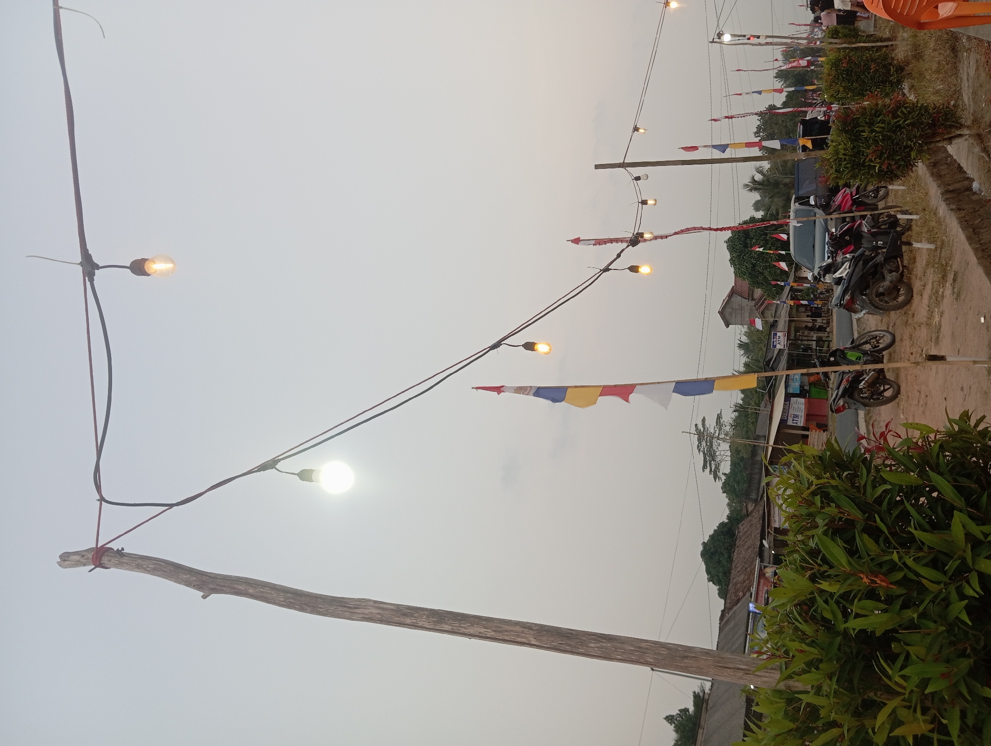 The image depicts an outdoor scene in Way Dente, Indonesia, likely during twilight. A wooden pole on the left supports a string of lights that extends across the frame, with several bulbs illuminated. A large green shrubbery is prominent in the foreground on the left, with red-tinged new growth. In the background, a street is visible with several small buildings, including one with an ATM sign. Various motorcycles are parked along the street. Several Indonesian flags, in red and white, are displayed on poles along the road. The sky is overcast, creating a muted light. The overall impression is that of a local gathering or street scene.