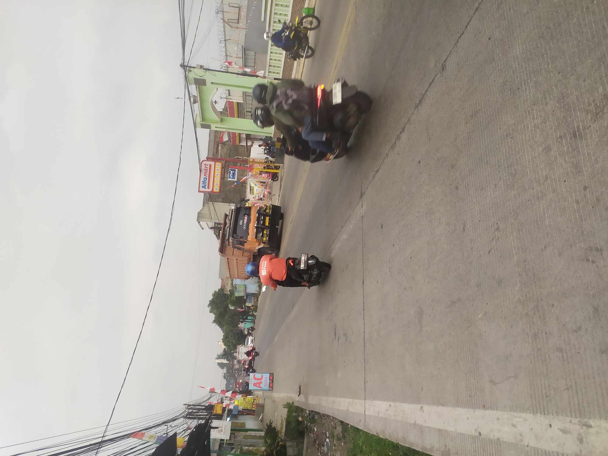The image depicts a street scene in Kragilan, Indonesia, on an overcast day. A gray, two-lane road runs through the center of the frame, with a concrete sidewalk on the left. Several vehicles are present: an orange dump truck is parked on the right side of the road, a man wearing an orange shirt and blue helmet is riding a black scooter in the middle of the road, and two people are on a dark-colored scooter traveling behind him. Further down the road, a person on a yellow motorcycle is visible on the right. Buildings line the left side of the street, including a store with an "Alfmart" sign and an "AC Mobil" advertisement. Red and white flags are displayed on poles along the roadside. The environment appears to be a commercial or residential area with utility wires crisscrossing overhead.