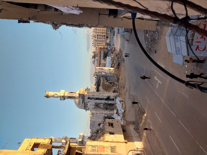 A desolate cityscape features a severely damaged mosque with its minaret prominently displayed against a clear sky. A broad street cuts through the scene, marked with a directional arrow and littered with debris. Several figures, some appearing as silhouettes, are scattered along the roadside and within the rubble. Heavily damaged buildings frame the vista, suggesting a scene of destruction and desolation. Electrical wires and fragments of structures hang precariously, adding to the atmosphere of ruin.