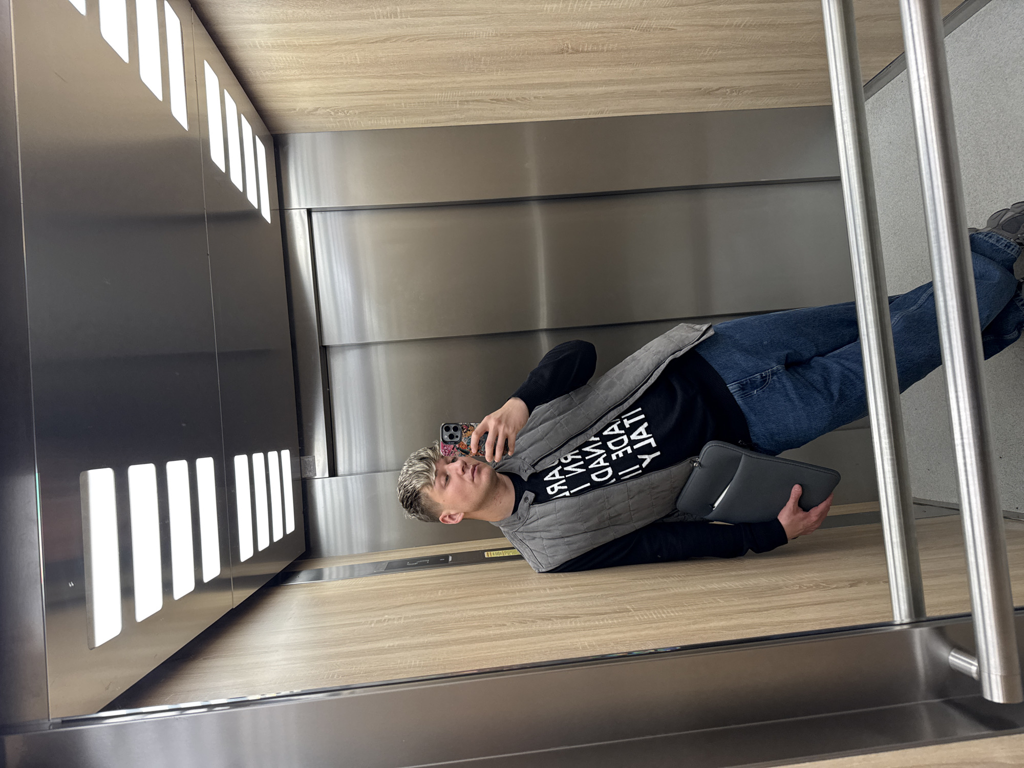 A young man is taking a selfie in an elevator. He is holding a phone in his right hand and a gray laptop case in his left. The man is wearing a black shirt with white lettering, a gray vest, and blue jeans. He has short, light-colored hair. The elevator has a wood-paneled floor and walls with horizontal metal panels. There are vertical rows of rectangular lights on the left wall of the elevator, and metal bars on the right, in front of a gray wall. The man’s location in Plovdiv, Bulgaria, cannot be determined from the image.