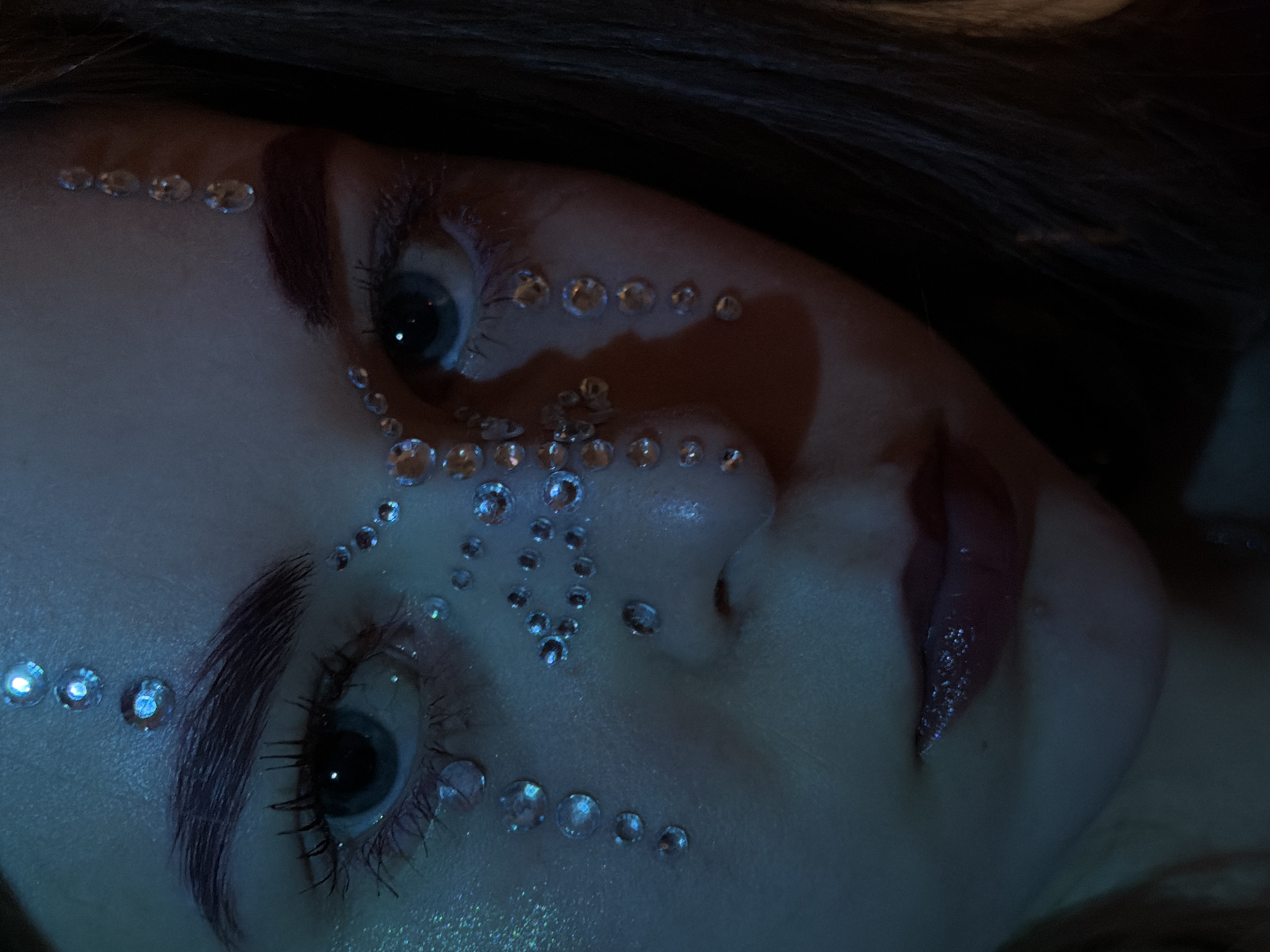 A close-up, medium shot features a person's face, decorated with small, reflective rhinestones. The rhinestones are arranged in patterns along the brow bone, under the eyes, and across the nose. The person has dark hair visible at the top and side of the frame. Their eyes are open, and they appear to be looking slightly upward. The lighting has a cool, blueish tint. The setting or location is not discernable in the image, and there is no information to suggest it is in Perm, Russia.