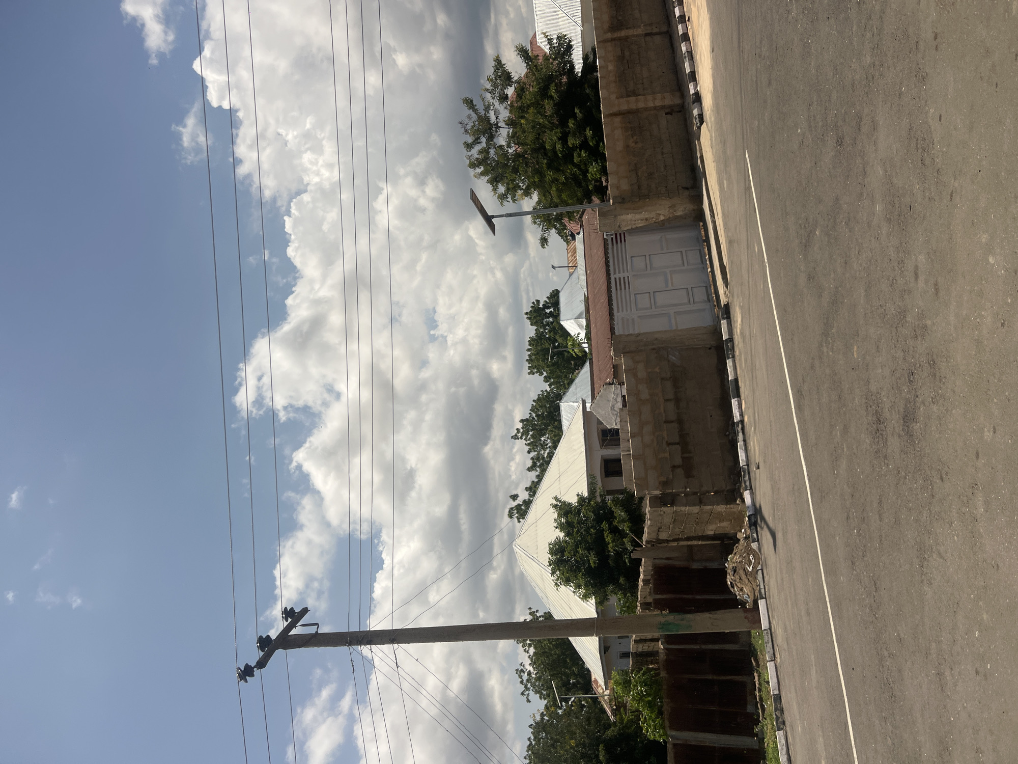 The image depicts a street scene in Chum, Nigeria, under a sky with significant cloud cover and visible patches of blue. In the foreground, a gray asphalt road extends across the lower portion of the frame, featuring a single solid white line painted along its surface. The road is bordered by curbs painted with alternating black and white stripes. On the left side of the road, a small pile of earth or debris is visible at the base of the curb.
A tall concrete utility pole with multiple dark insulators and several horizontal power lines dominates the left side of the midground. Behind the road and extending from left to right, a low wall constructed from unpainted concrete blocks encloses properties. A light-colored, likely white, double-panel metal gate is integrated into this wall on the right side.
Behind the wall and amidst dense green foliage, several residential structures are visible. A prominent building in the center-left features a large, light-colored, multi-pitched or pyramidal roof, likely made of corrugated metal. Other houses with similar metal roofs, varying in color from silver to reddish-brown, are partially visible through the trees. On the far right, a solar-powered street light mounted on a pole stands adjacent to a tree, with its solar panel oriented upwards. No individuals, explicit actions, or interactions are present in the scene.
