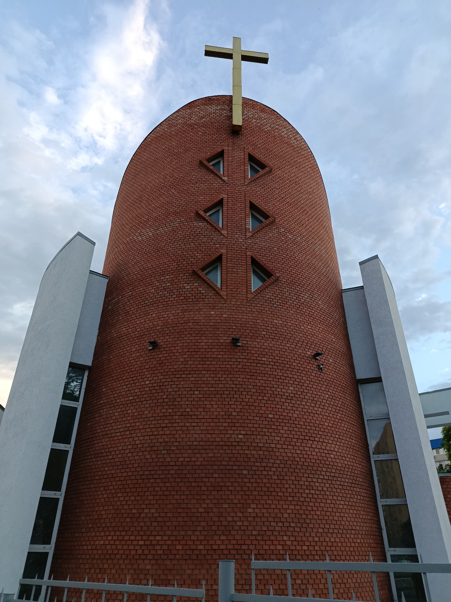 The image shows a low-angle, full-frame exterior shot of a modern church structure, possibly St. Peter and Paul Cathedral, located in Saratov, Russia. The building features a prominent, curved red brick facade topped with a golden cross. Several triangular windows are arranged in a diamond pattern on the brick wall. On either side of the central brick structure are white, angular architectural elements with vertical windows. The sky above is partly cloudy. In the foreground, a metal fence is visible. There are no people or animals present in the image.