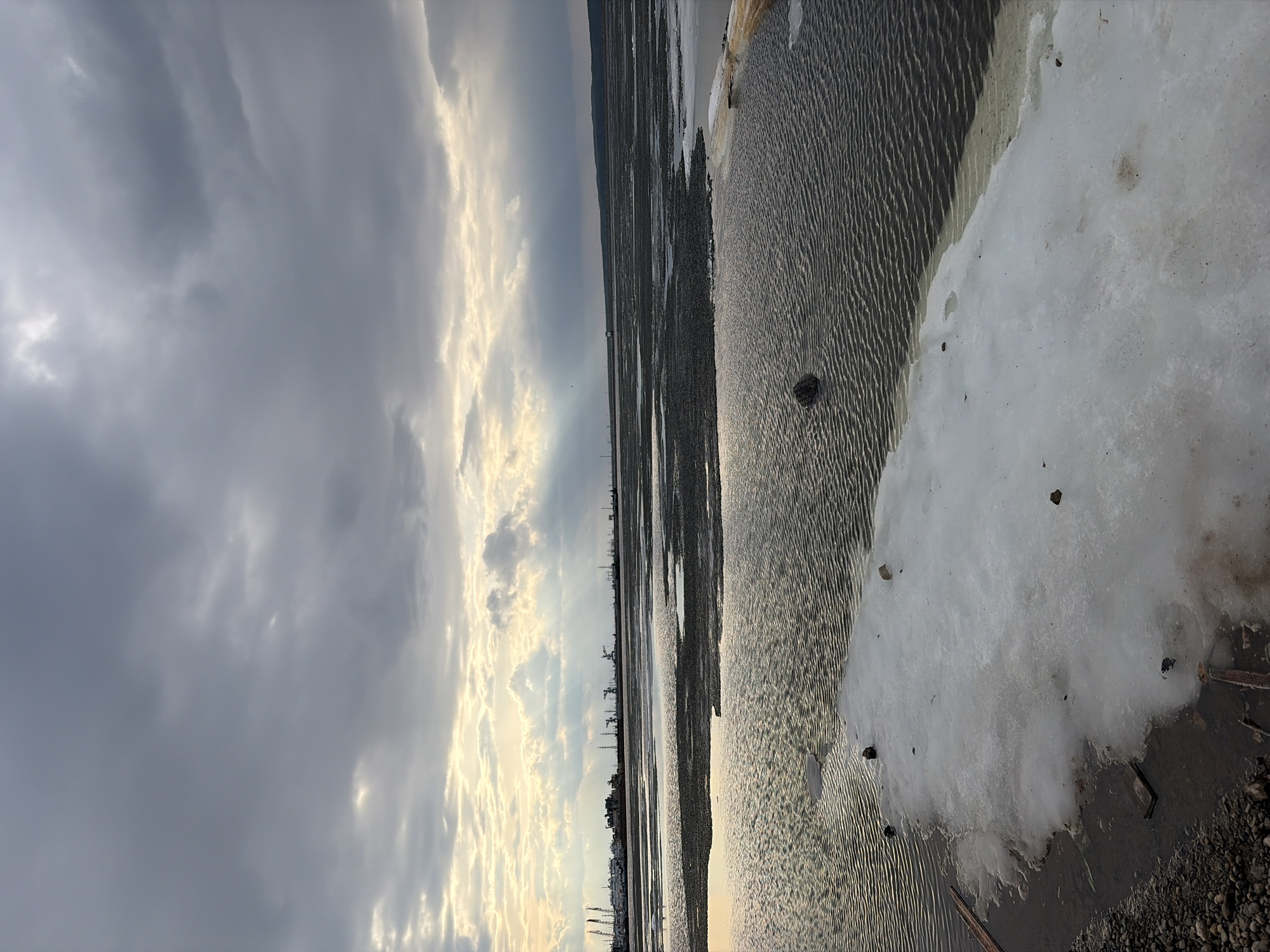 The image depicts a landscape in Chaikovsky, Russia. The foreground shows a muddy beach covered with shallow water. The water's surface is rippled, suggesting a slight breeze. Patches of melting snow and ice are visible near the edge of the beach, indicating a transitional season. In the distance, a cityscape with tall structures like industrial cranes and buildings is silhouetted against the horizon. The sky is overcast with dark, heavy clouds tinged with lighter hues of gold and white where the sun is breaking through. The light in the scene is subdued, contributing to a somewhat bleak and cold atmosphere.