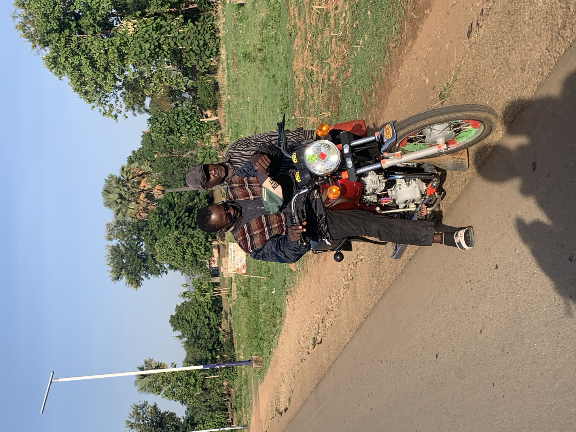 Two African men are seated on a red motorcycle parked on the side of a paved road. The man in the front wears a plaid shirt and black pants, with white and black footwear. The man behind him wears a striped shirt and a cap. The motorcycle has red, green, and silver decorations. The location is Maigizo, Nigeria, as indicated by a sign in the background. The environment includes trees, grass, and a blue sky. A signpost is visible on the left.