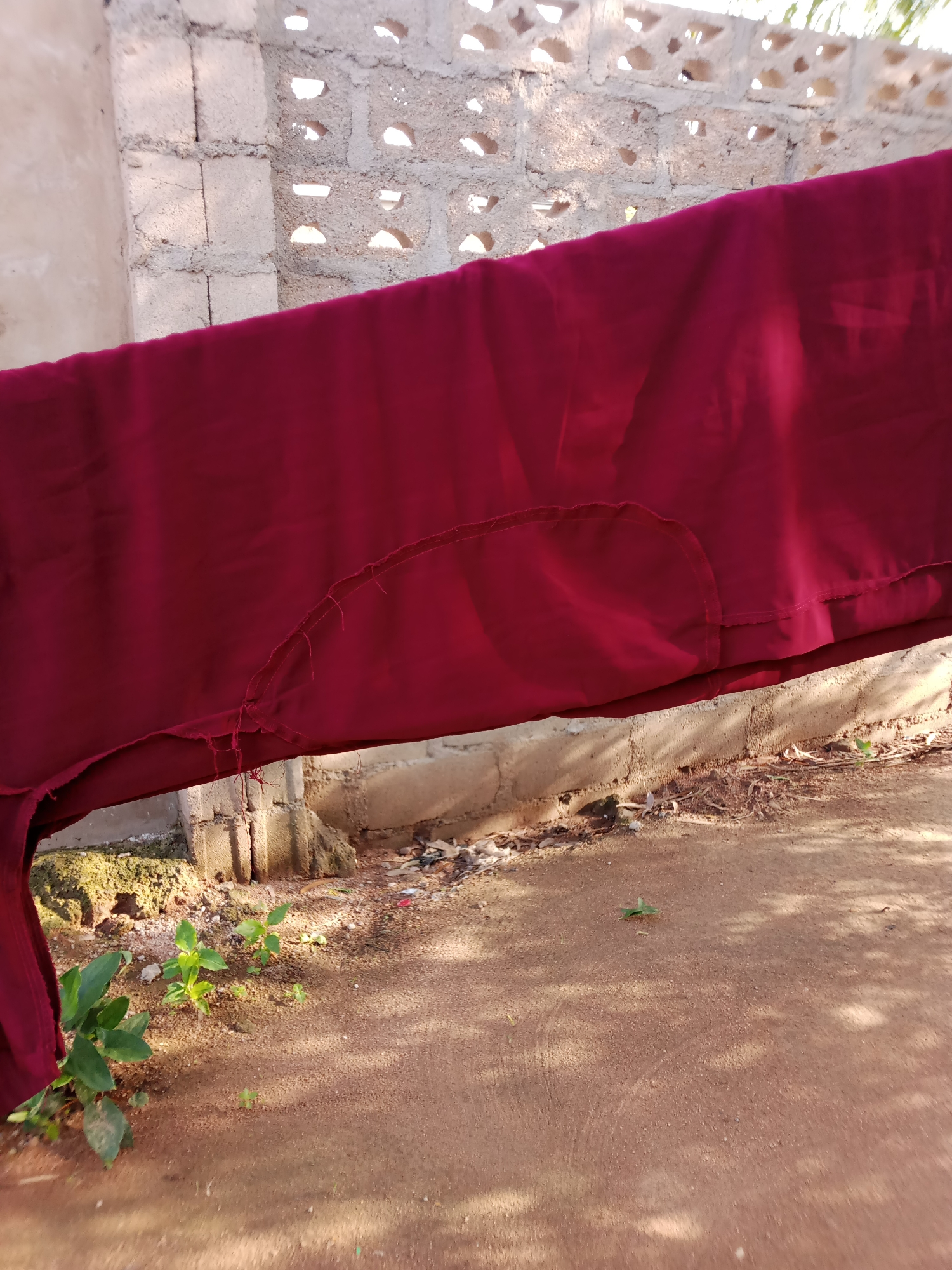 The image depicts a close-up view of a dark red fabric, possibly a garment or cloth, hanging from a line. The fabric appears damaged along its lower edge, with visible fraying and loose threads. In the background, a wall constructed of concrete blocks is visible, with some blocks having decorative openings. The ground in the foreground is reddish-brown dirt, with scattered debris and some small green plants. The overall scene suggests an outdoor setting, possibly a yard or outdoor drying area, in Jauro Yinu, Nigeria. The lighting indicates it is daytime, with shadows cast on the ground, suggesting sunlight. No people are present in the image, and the focus is on the fabric and its immediate surroundings.