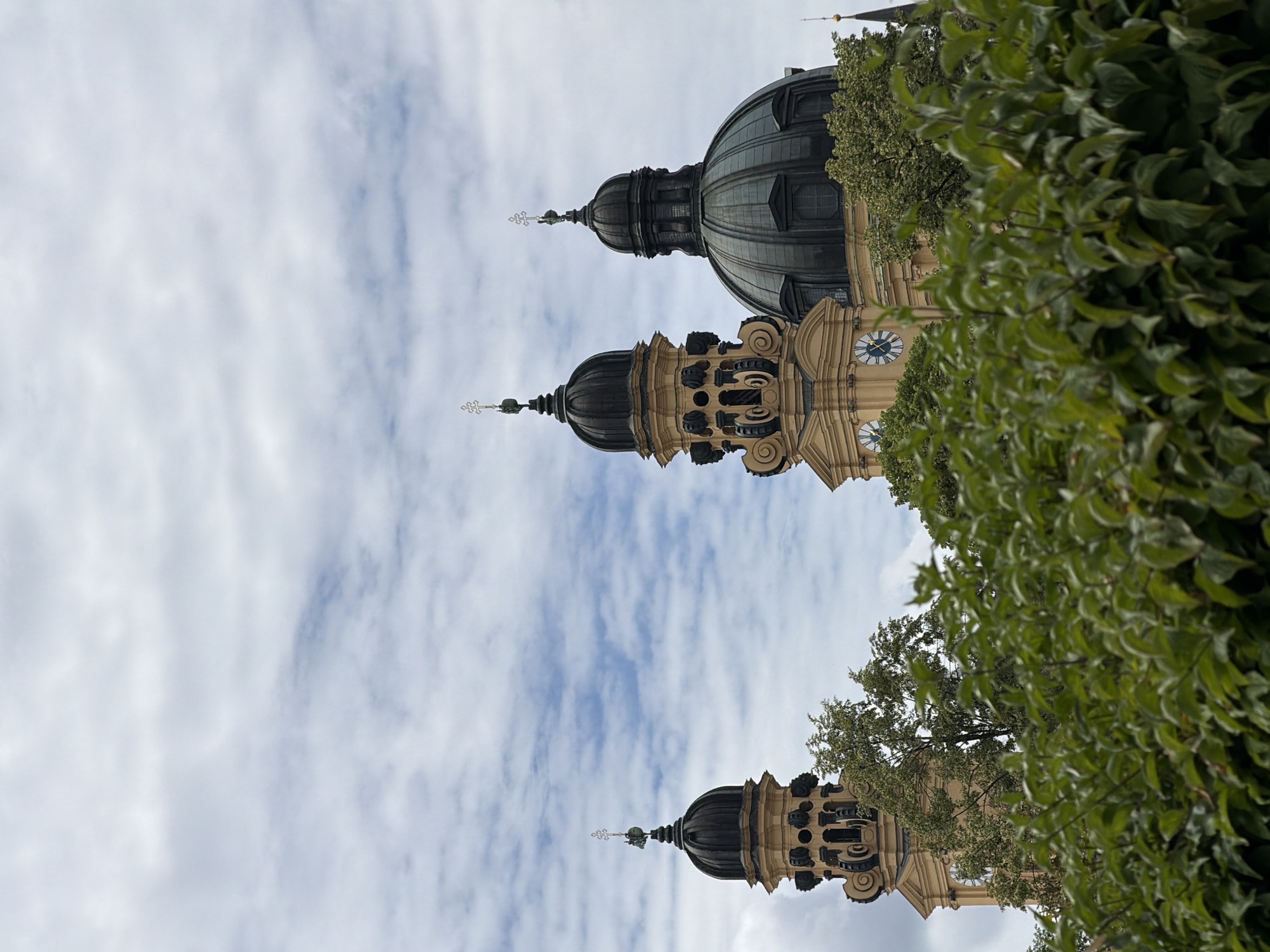 The image shows a view of the towers and domes of a large building, likely a church or cathedral, in Munich, Germany. The architecture features light yellow stone and dark green domes, topped with crosses. Two prominent towers with rounded tops are visible on the left, and a larger dome is visible to the right. Clocks are visible on the faces of the towers. Lush green foliage is in the foreground, obscuring the lower part of the structure. The sky above is a mix of white and blue clouds. The scene is tranquil and architectural.
