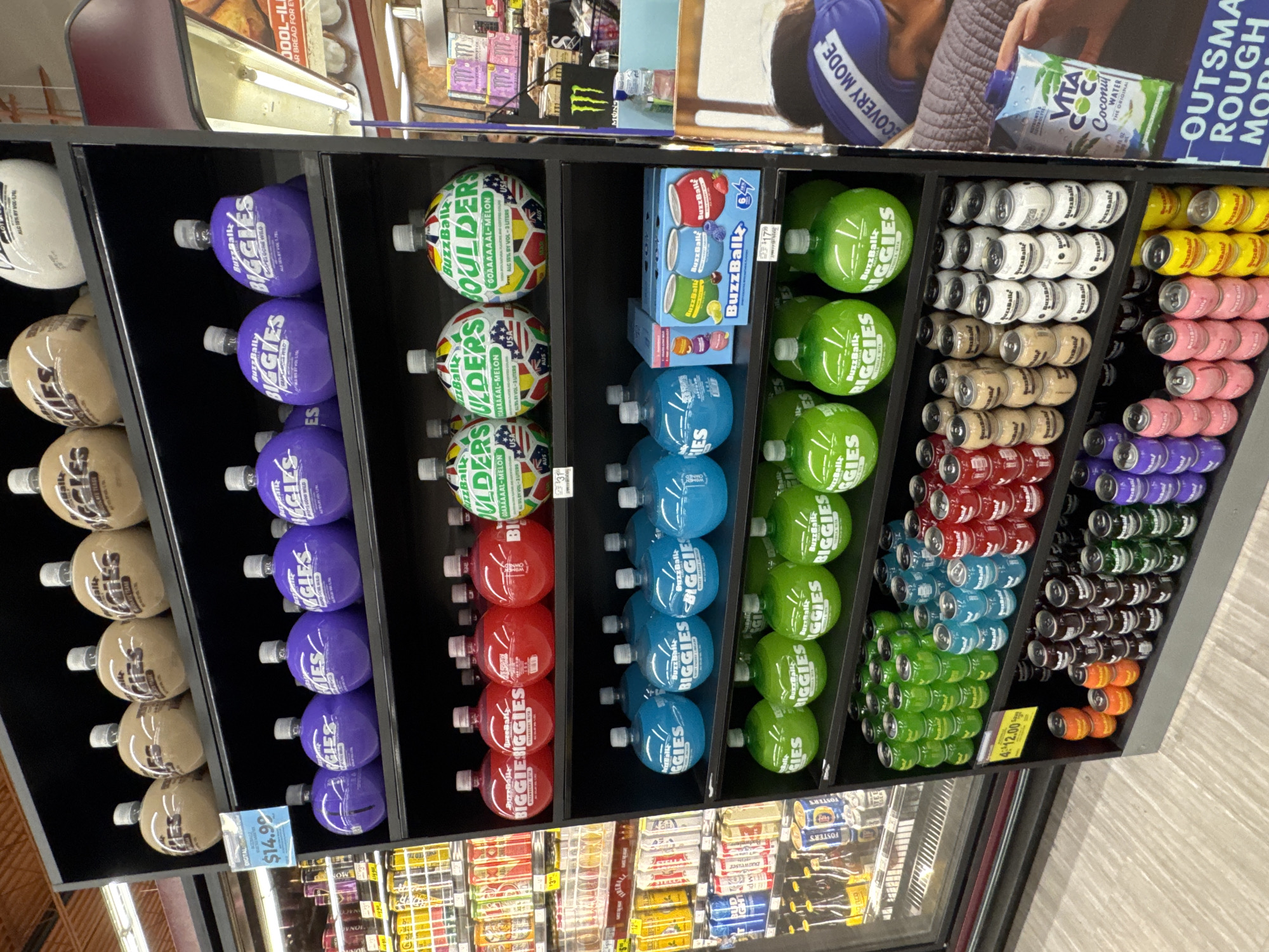 This image captures a brightly lit retail display of alcoholic beverages, likely within a supermarket or liquor store in Glendale Heights, USA. The scene showcases a variety of BuzzBallz brand products, neatly arranged on black shelving units.
The primary subjects are rows of BuzzBallz "BIGGIES" and "BOULDERS" bottled beverages, along with numerous BuzzBallz canned drinks. On the left, several large, spherical bottles of "BuzzBallz BIGGIES" are displayed vertically, appearing in colors such as brown, purple (labeled "GRAPE GONE WILD"), red, blue, and green. Some brown bottles are associated with "Chocolate" or "Vanilla" flavors. In the center, "BuzzBallz BOULDERS" are stacked horizontally, recognizable by their colorful, patterned spherical containers, with visible text indicating "GOAAAAAAL-MELON" flavor and "ALC 15% BY VOL - 3 LITERS." A small multi-pack of BuzzBallz featuring Strawberry, Blueberry, Lemon, and Lime flavors is also present.
To the right, numerous small, spherical BuzzBallz cans are stacked horizontally in various colors, including white, brown, red, blue, green, yellow, pink, and purple, suggesting a wide array of flavors. A yellow price tag reading "4-12.00" is visible beneath some of the canned products. Another digital price tag, "$14.99," is seen near the purple "BIGGIES."
Above the product display, a promotional poster or advertisement features a person wearing a "RECOVERY MODE" hat and holding a "VITA COCO Coconut WATER," with text also visible reading "OUTSMART ROUGH MORNING." Other visible text includes "MONSTER" and "FOSTER'S" on products in the refrigerator unit below the main display. The setting is indoors, and no specific time of day, weather, or human emotions are discernible.