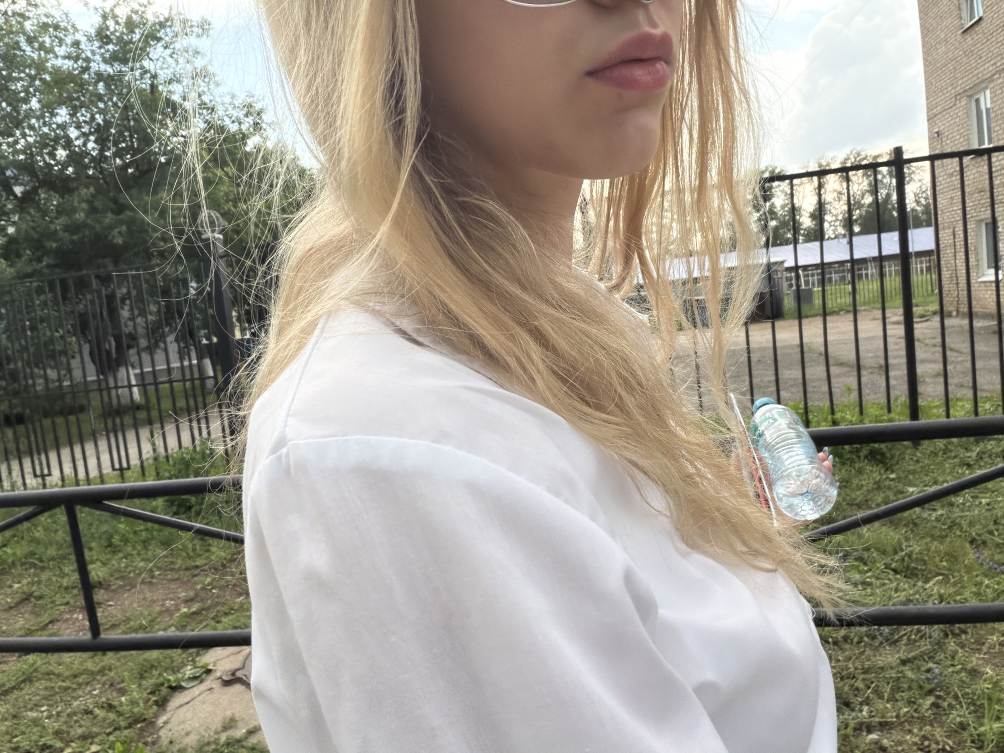 A young Caucasian woman with blonde hair stands outdoors, her face partially visible from the side. She is wearing a white shirt and holding a clear plastic water bottle in her right hand. The background consists of a black metal fence, green grass, trees, and a brick building. The sky above is partly cloudy.