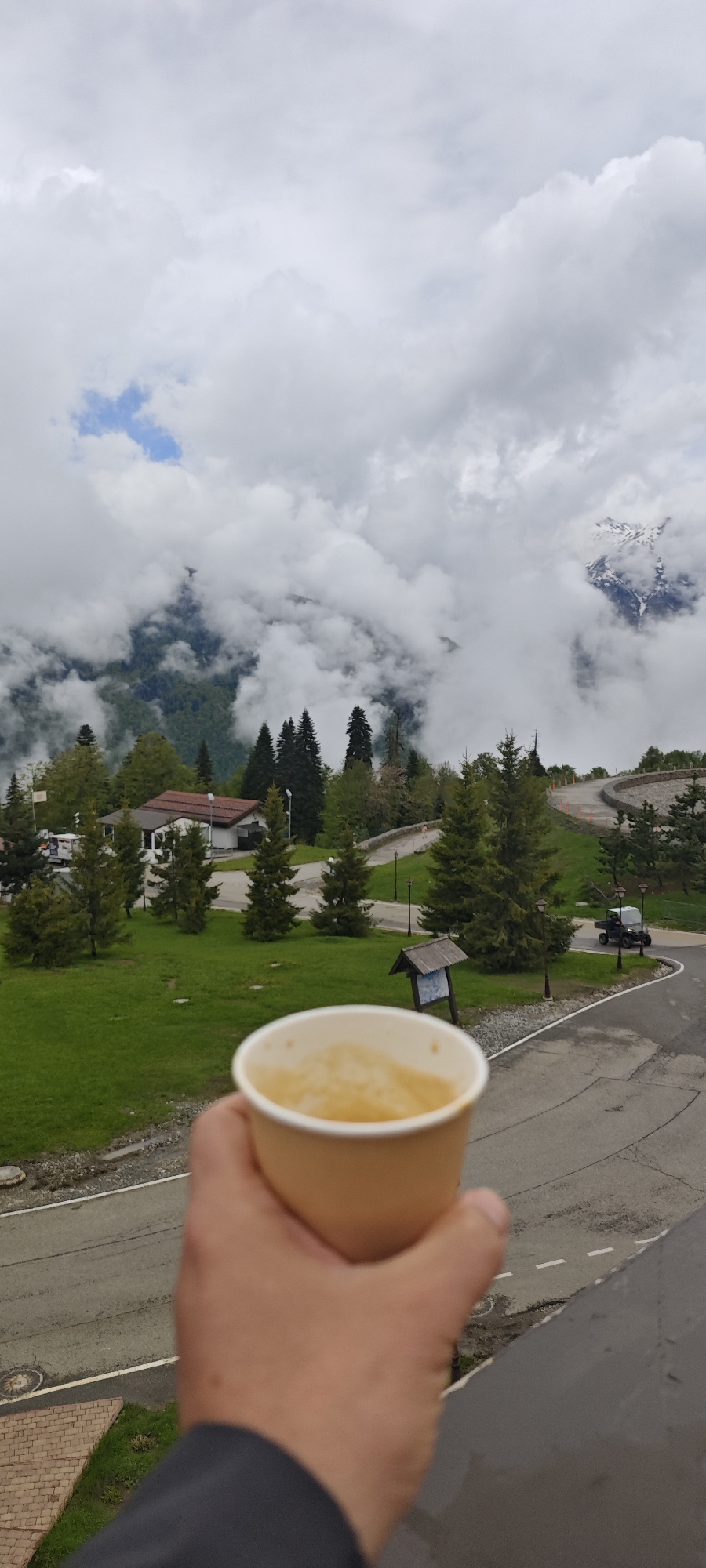A person holds a tan paper cup in their left hand against a backdrop of a mountainous landscape obscured by clouds, potentially in the area of Sochi, Russia. The cup appears to contain a yellow liquid, possibly tea or juice. The middle ground reveals a green, grassy area with a small building with a red roof, interspersed with evergreen trees. A road curves through the landscape, where a vehicle that looks like a golf cart is visible on the right side of the image. The sky is overcast, with clouds hanging low over the mountains. The image is shot from a slightly elevated position, possibly from a balcony. The foreground shows a section of what appears to be a grey stone or concrete structure.