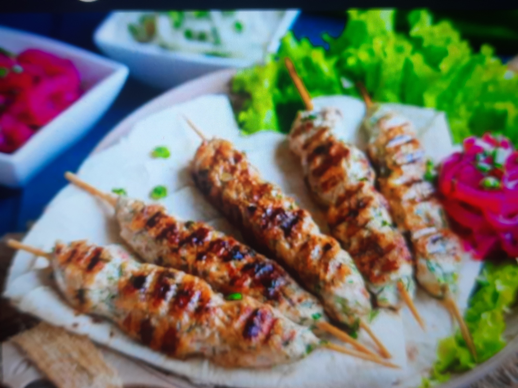 A plate of grilled chicken skewers served with flatbread, lettuce, and pickled onions is presented. The skewers are arranged on a white cloth, with grilled marks visible on the meat. The pickled onions are a vibrant pink, and the lettuce is a bright green. The image appears to be taken in a restaurant or a food establishment in Rosenheim, Germany, though no specific location is identifiable within the frame. The lighting is bright, suggesting it was taken during the day or in a well-lit indoor setting. There are no people or discernible interactions visible in the image.