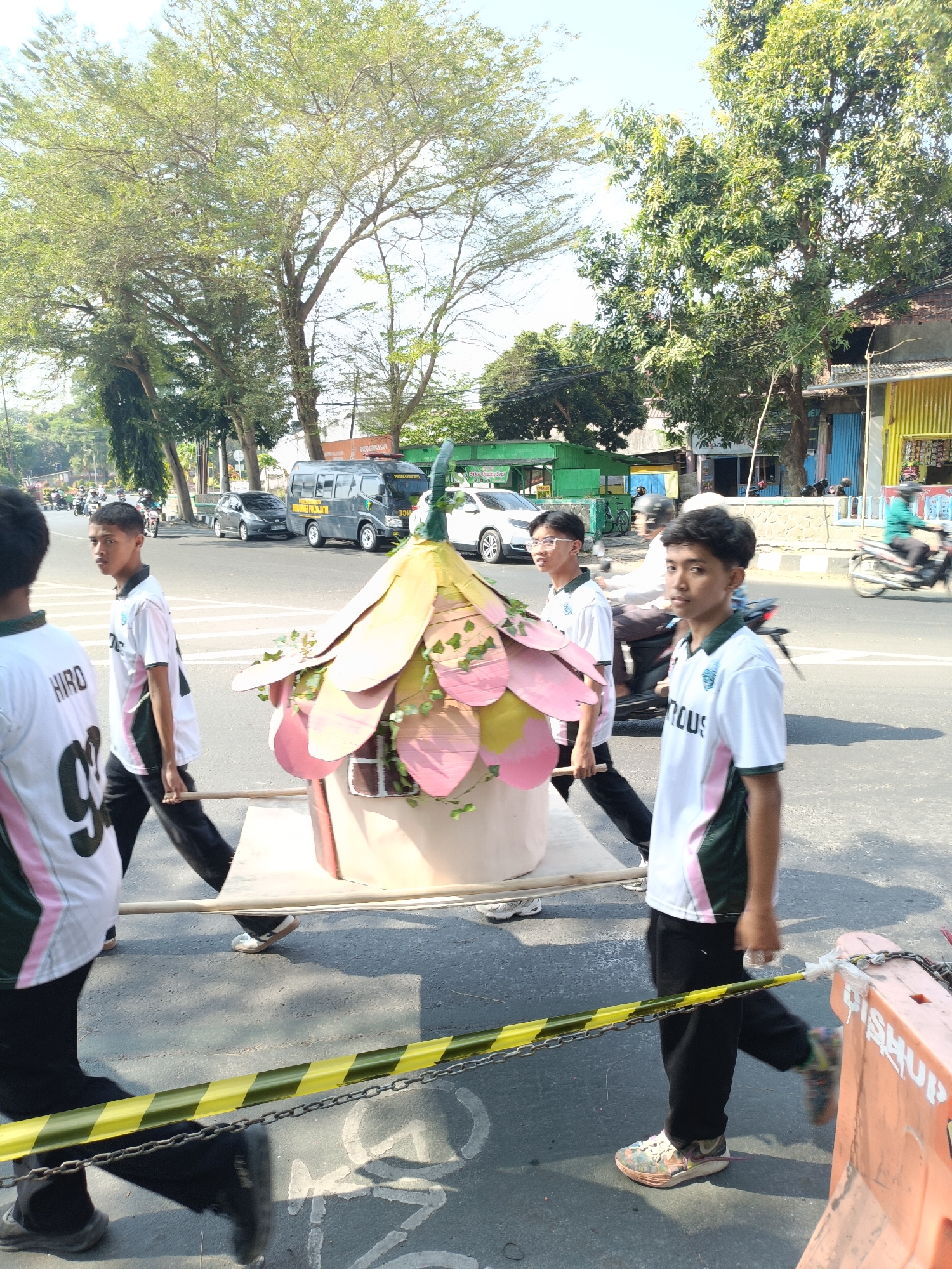 Four young males are walking across a street, carrying a decorative float shaped like a house with flower petals on its roof. The float is mounted on poles for carrying. The individuals are wearing white t-shirts with green and pink accents, and black pants. One person in the foreground has "HIRO" and "99" on their t-shirt. The scene is on a street in Kediri City, Indonesia, with trees lining the road, buildings in the background, and other vehicles on the street including a gray van and cars. There is a yellow and black striped caution tape barrier in the foreground and an orange traffic barrier on the right side of the image. The sky is clear and bright, indicating daylight.