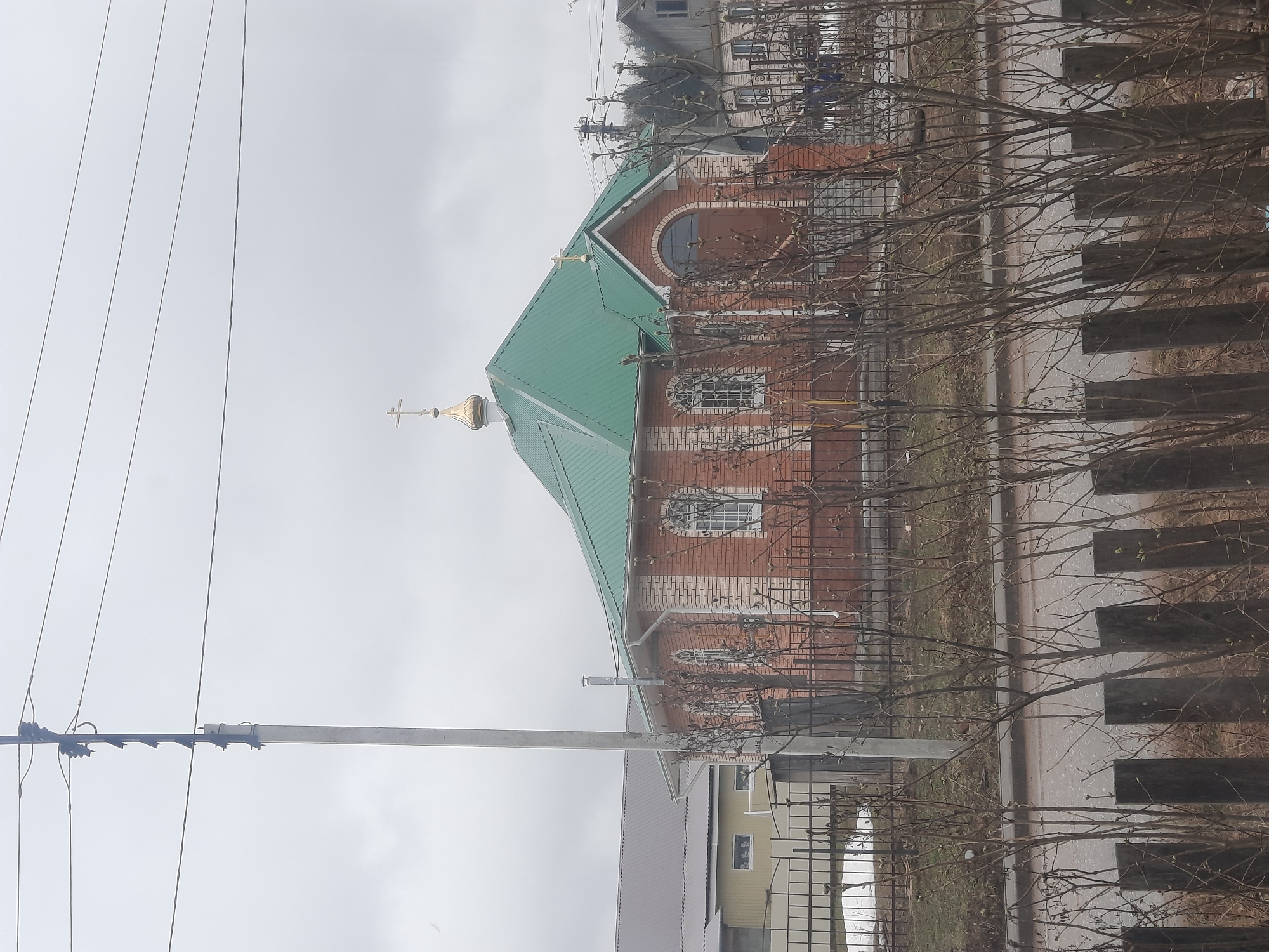 The image shows a Russian Orthodox church located in the city of Krasnogorskoye, Russia. The church is constructed of red brick with white brick accents, and it has a distinctive green roof. A golden onion dome topped with a cross sits atop the roof. The church is surrounded by a black metal fence. In the foreground, there is a row of bare trees and a wooden fence. A utility pole with power lines is visible to the left of the church. A gray sky suggests overcast weather conditions. The architecture and religious symbols indicate the cultural context of the location.