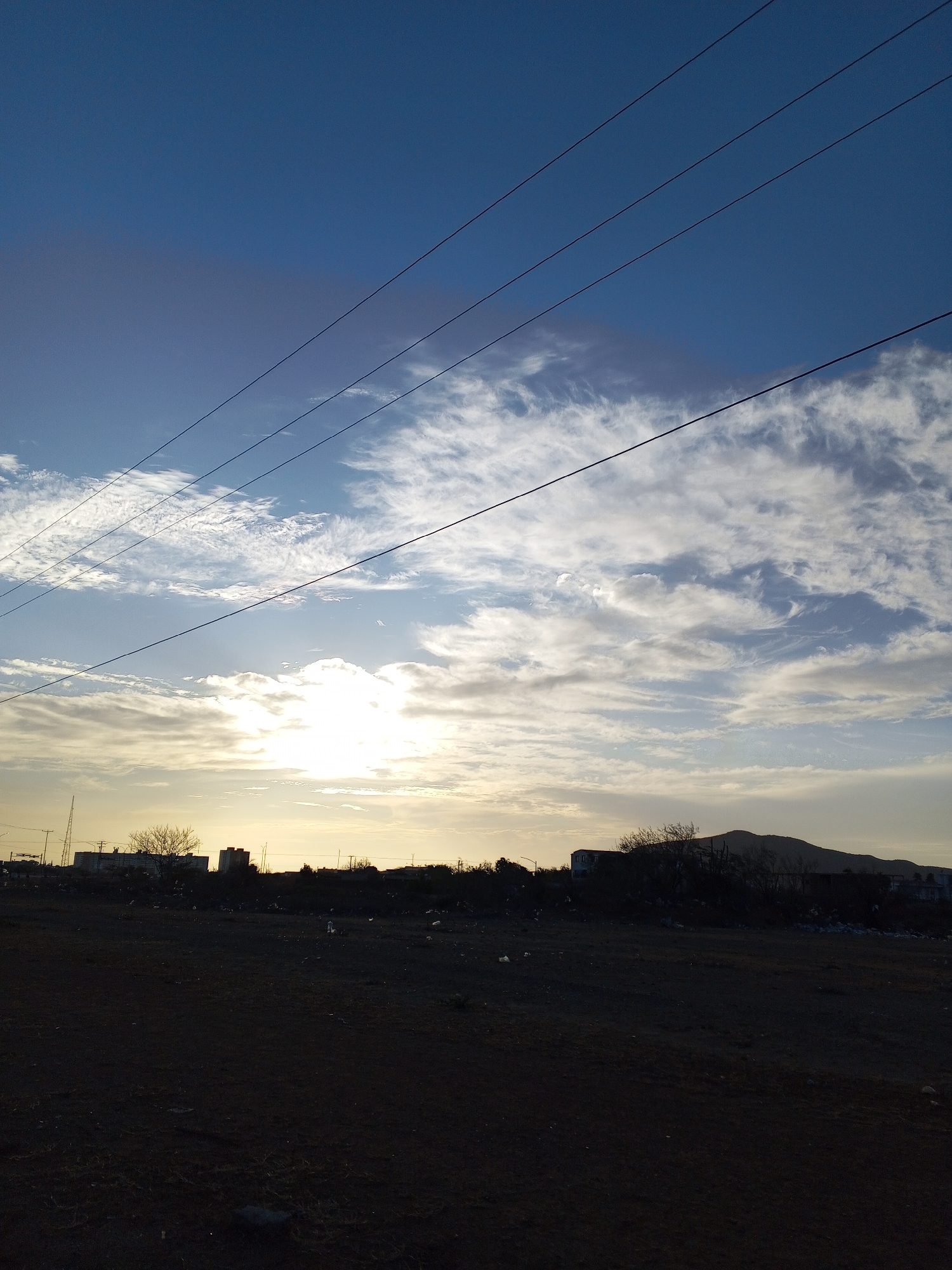 The image depicts a landscape at dusk or dawn in an urban area of Altagracia, Venezuela. The foreground is dominated by a dark, dirt field. In the midground, a silhouette of buildings and infrastructure including trees, houses and a mast are visible. A low hill appears on the right. Above, the sky is blue with scattered white clouds and electrical wires traverse the scene horizontally. The sun is obscured behind the horizon, creating a bright glare.